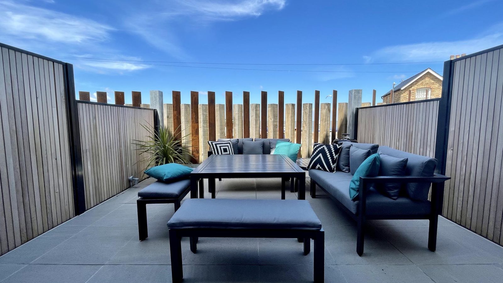 A modern outdoor terrace, known as The Look Out, features a black table flanked by two black benches with blue cushions and a matching black sofa adorned with patterned and solid blue pillows. The space is enclosed by wooden fencing, and the sky is bright with a few clouds.