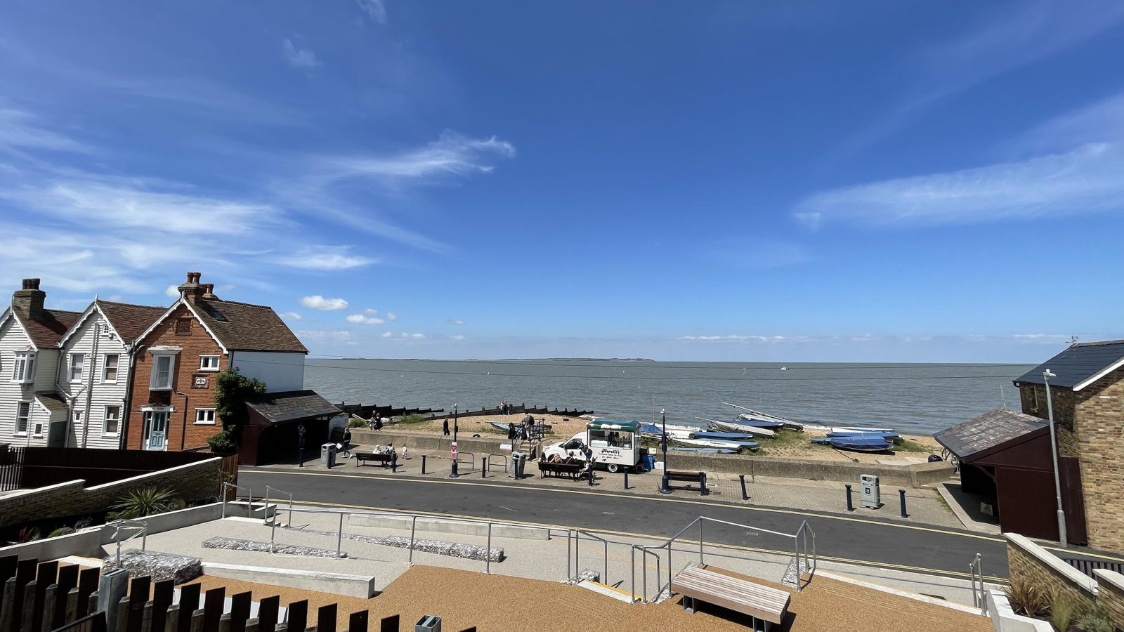 A scenic view of The Look Out seaside village with a clear blue sky. The image shows a beach area with a few boats on the shore. There are houses and a paved walkway overlooking the waterfront. The horizon meets the calm sea, and light clouds are scattered in the sky.