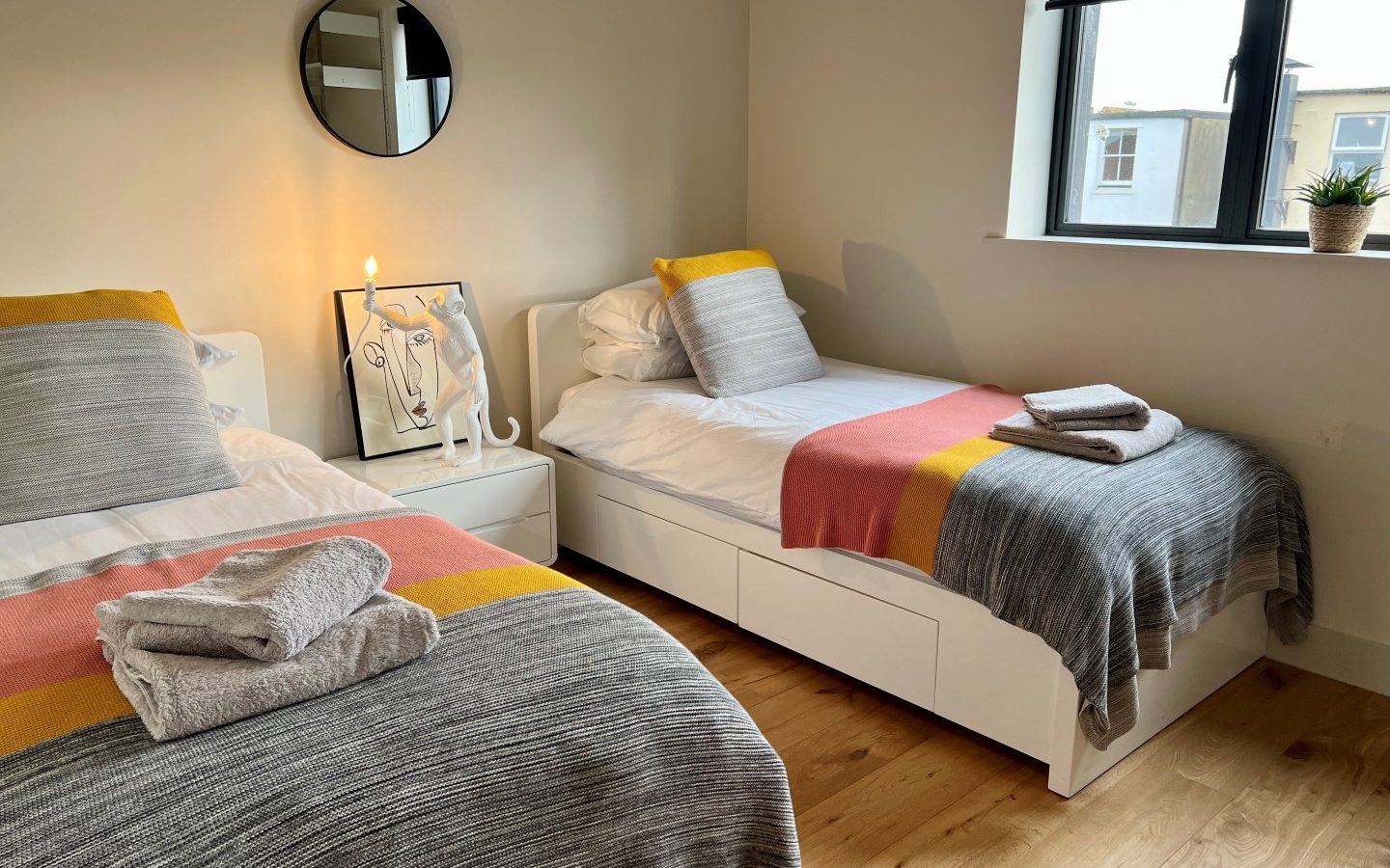 A cozy bedroom at The Look Out features two white single beds with gray, pink, and yellow striped blankets and neatly folded towels on top. A bedside table with a decorative lamp and framed art sits between the beds, with a round mirror above it. A window with a dark blind and small potted plant allows natural light into the room, highlighting the wooden floor.