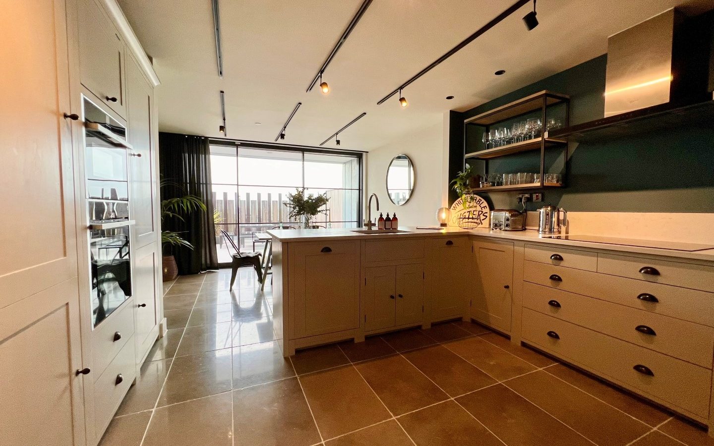 A spacious, modern kitchen with white cabinetry, a central island, and dark countertops. Stainless steel appliances, open shelving, and potted plants add to the décor. Large windows and glass doors look out to a balcony, allowing ample natural light in.