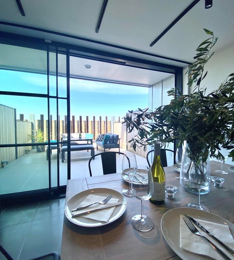 A modern dining area with a wooden table set for two—featuring white plates, silver cutlery, an empty wine glass, and a bottle of wine. In the background, The Look Out boasts a balcony with outdoor seating and a glass railing, offering a stunning view of the sky and horizon.