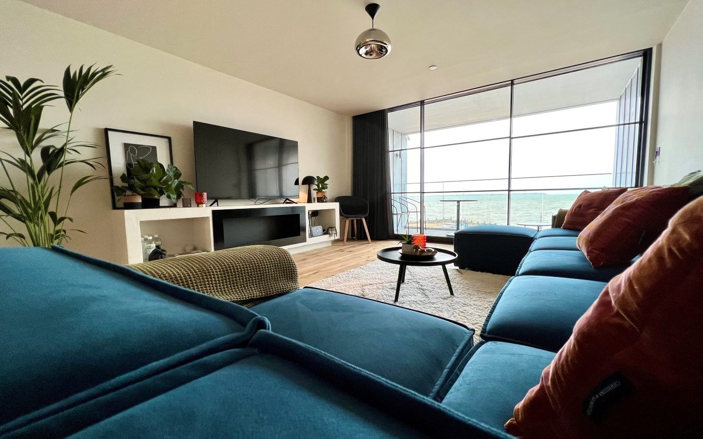 Modern living room with a large blue sectional sofa, colorful pillows, and a round coffee table. A large flat-screen TV is mounted on a white media console, while tall glass doors open to 