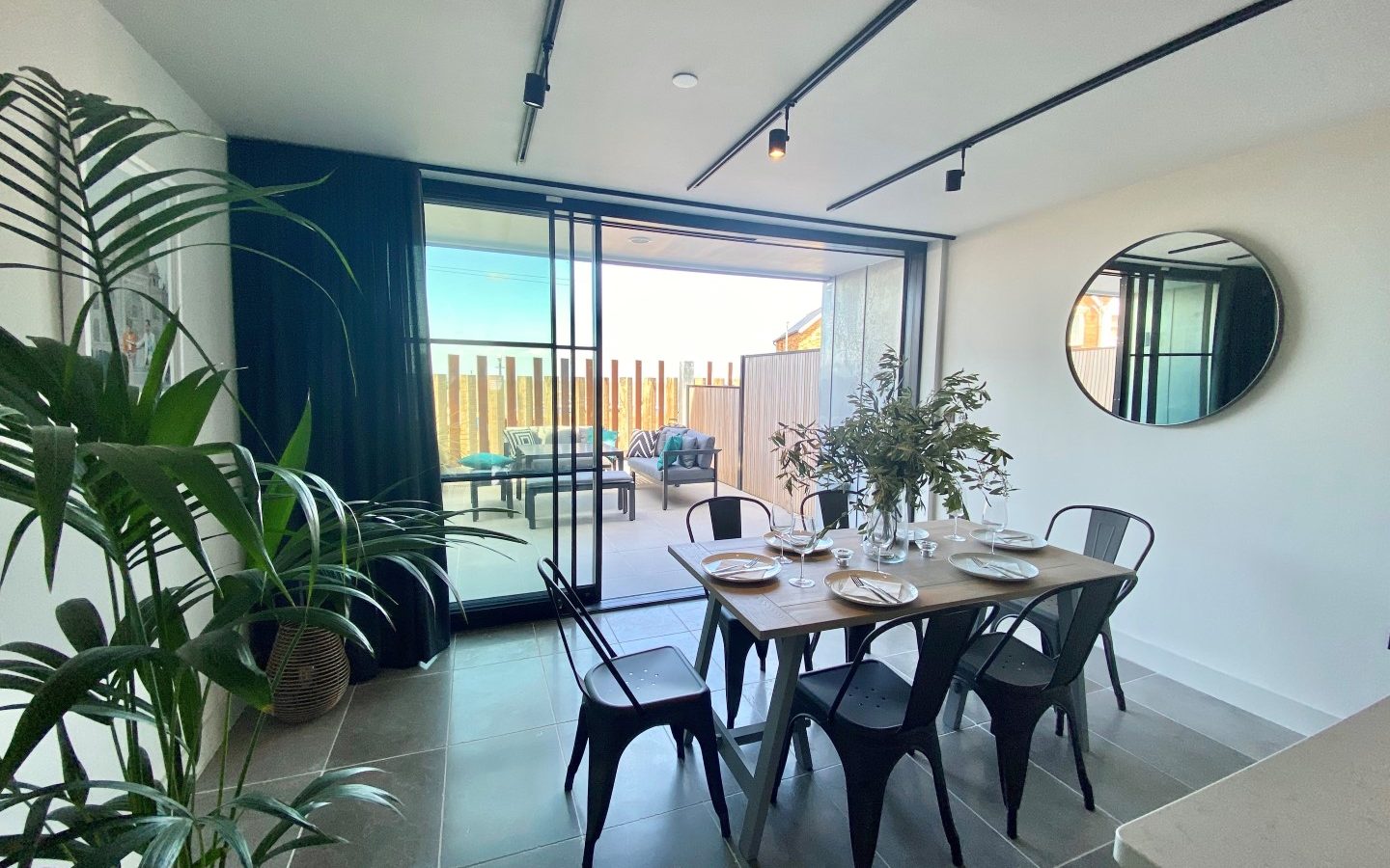 The Look Out features a modern dining area with a rectangular table set for six, black chairs, and a large mirror on the wall. Glass sliding doors open to a patio furnished with outdoor seating. There's a tall plant on the left and track lighting on the ceiling, enhancing the sophisticated ambiance.