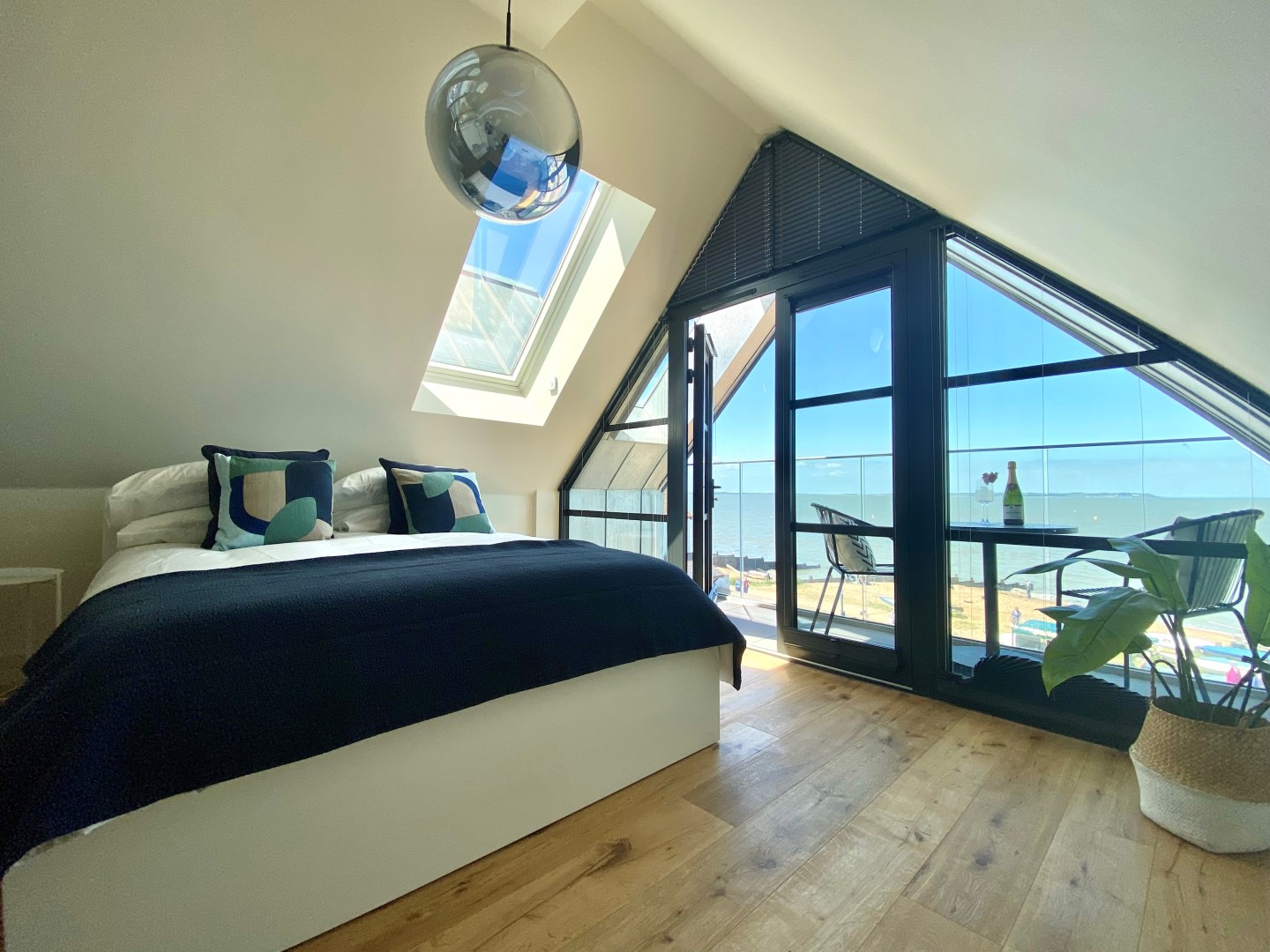 A modern bedroom with a slanted ceiling features a bed with black and white bedding and colourful pillows. A large window and glass door lead to a balcony with a table and chairs, providing a scenic view of the beach and ocean. A hanging spherical light fixture adds ambiance. Ideal seaside accommodation near London