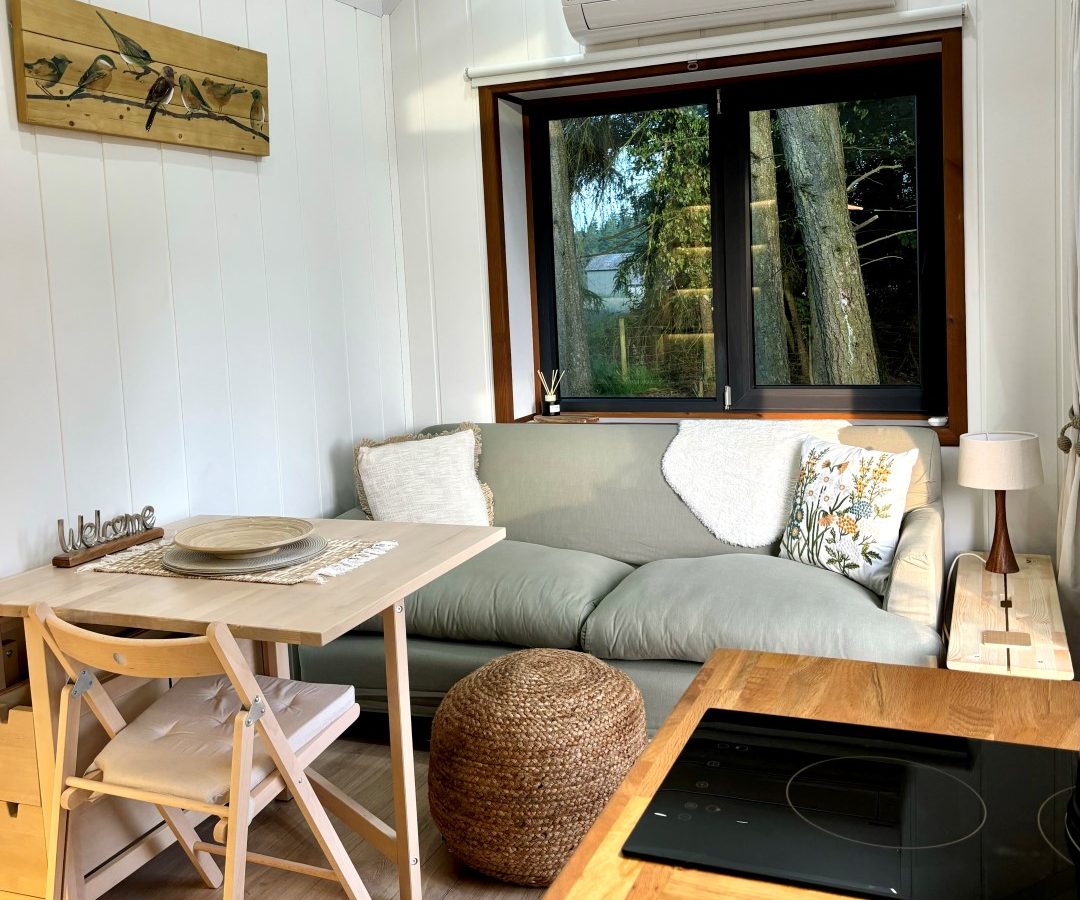 A cozy living area in a tiny house features a small dining table and chairs, a comfortable sofa, and a pouf. With white paneled walls, a large window reveals stunning woods outside and includes an electric cooktop in the foreground. A charming wooden decor piece completes the inviting space.