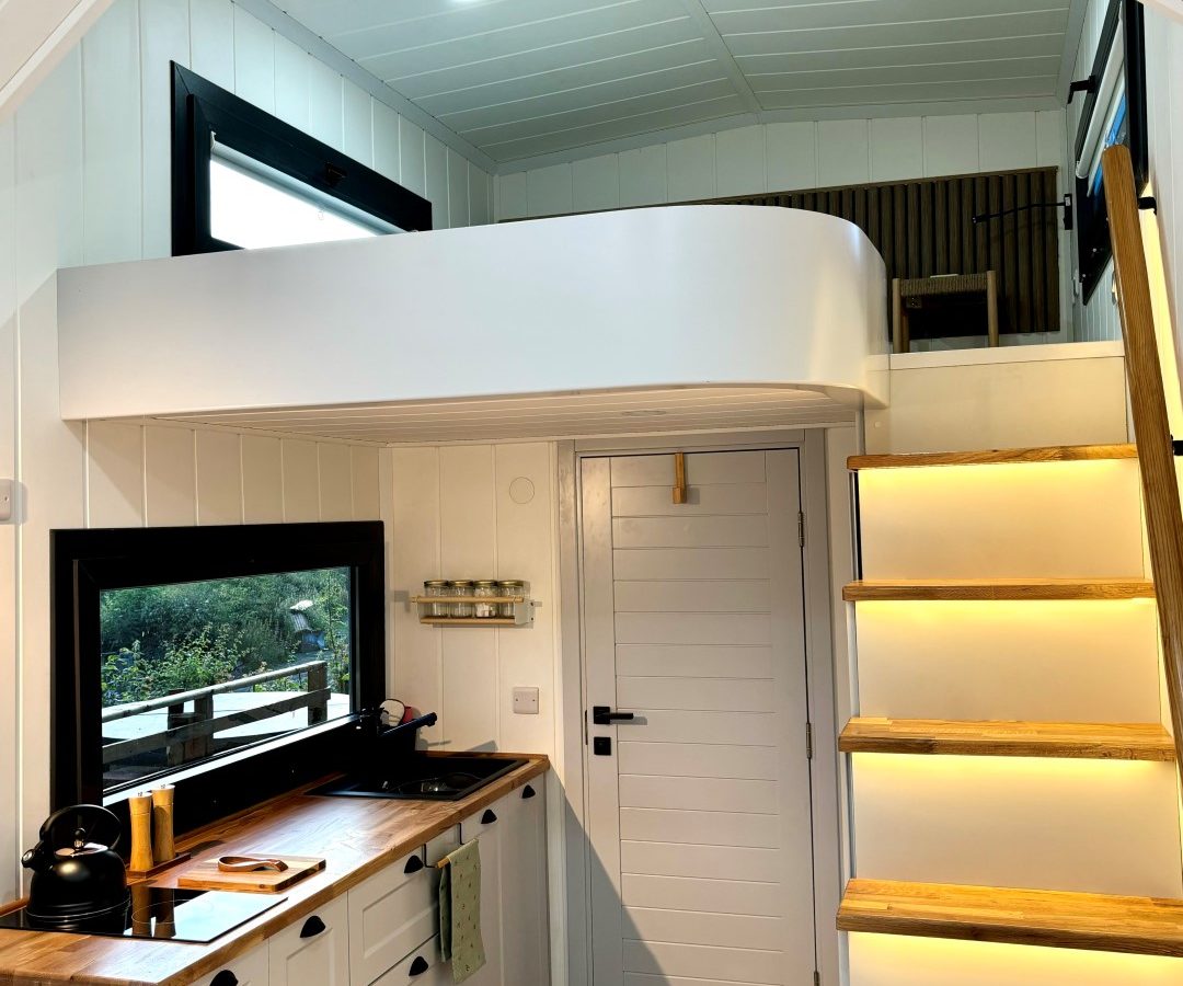 A modern tiny house interior showcases a cozy kitchen with light wood countertops and white cabinetry. A staircase with built-in lighting ascends to an elevated sleeping loft, while large windows offer ample natural light and views of the surrounding woods.