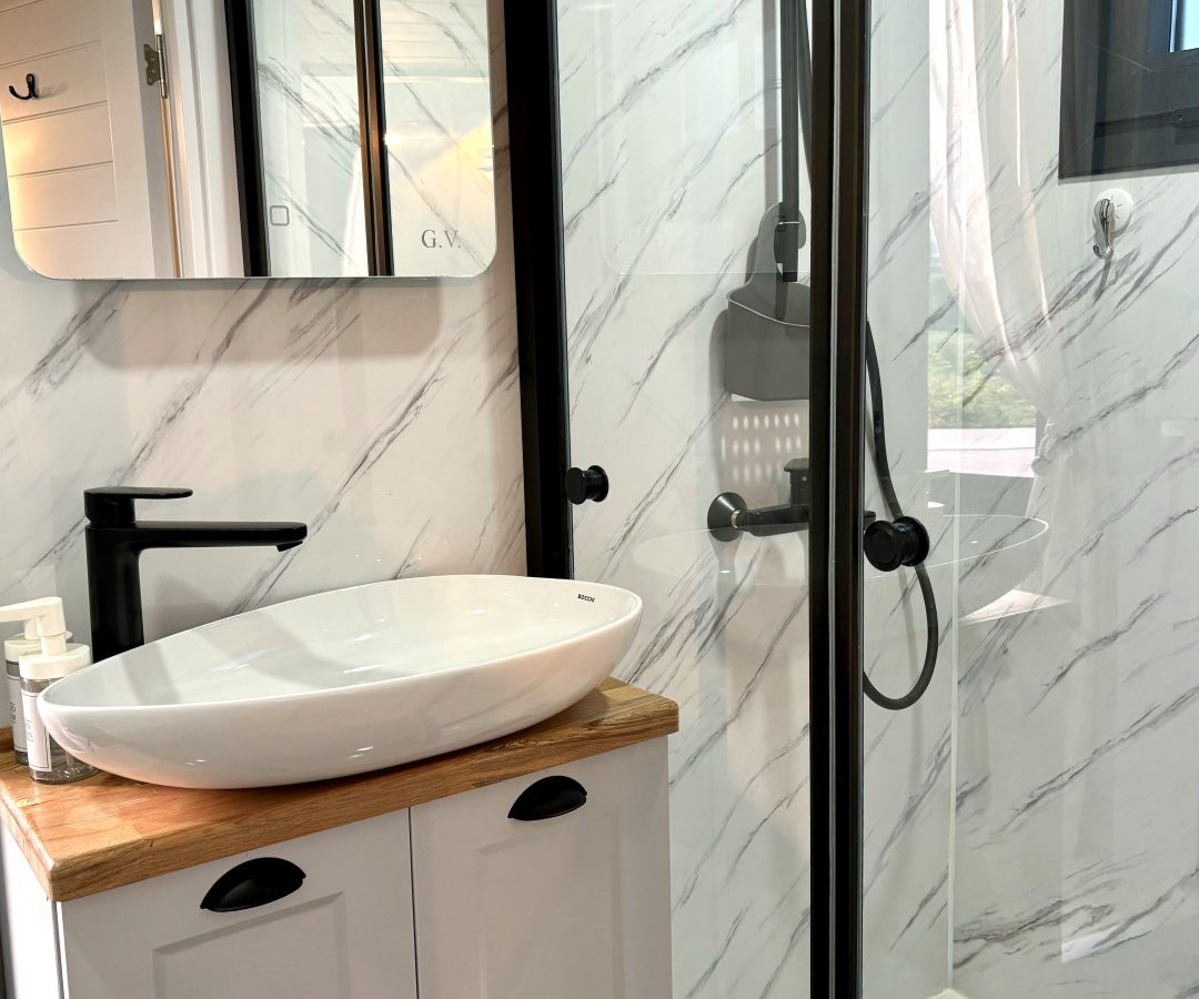 A modern bathroom in a tiny house features a glass-enclosed shower with a rainfall showerhead. A white sink sits on a wooden countertop above a white cabinet. The walls have a marble-like finish, and a sleek black faucet complements the design nestled in the woods.