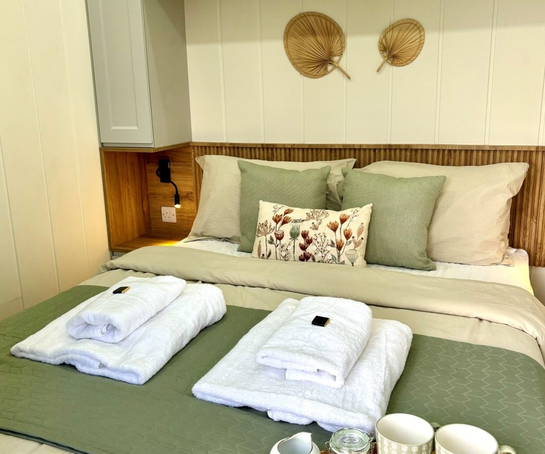 A cozy bedroom in a tiny house in the woods, with a neatly made bed featuring green and beige bedding. Two folded white towels sit on the bed, alongside a wooden tray holding two mugs, tea packets, and a glass jar. Decorative items and pillows add a touch of style to this charming retreat.