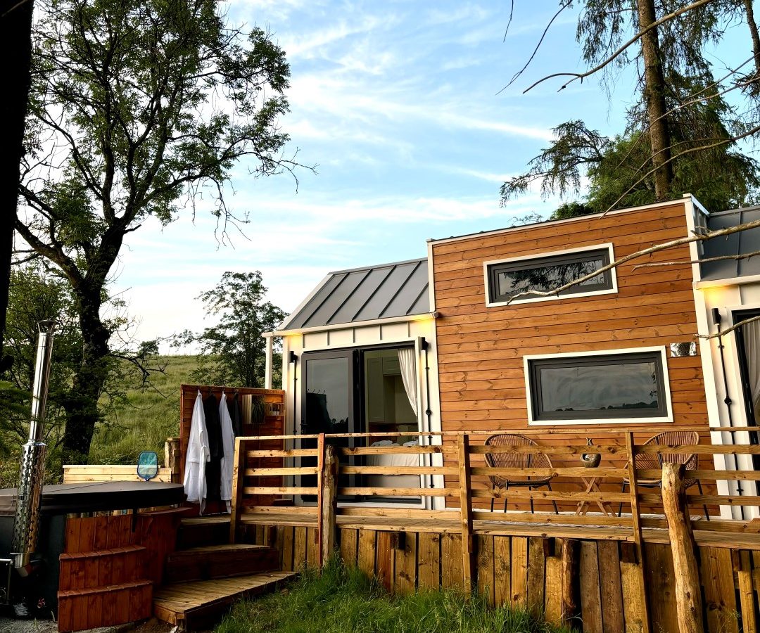 Nestled in the woods, this modern tiny house boasts large windows and a wooden exterior. A charming deck features a wooden hot tub and lounge chairs under a clear blue sky. A serene pathway winds toward the entrance, crafting an inviting outdoor scene among the towering trees.