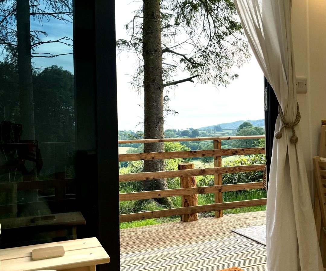 A cozy tiny house interior features an open door leading to a wooden deck, offering a scenic view of the woods and distant hills. White curtains frame the door, while inside, a small table and sofa invite relaxation.