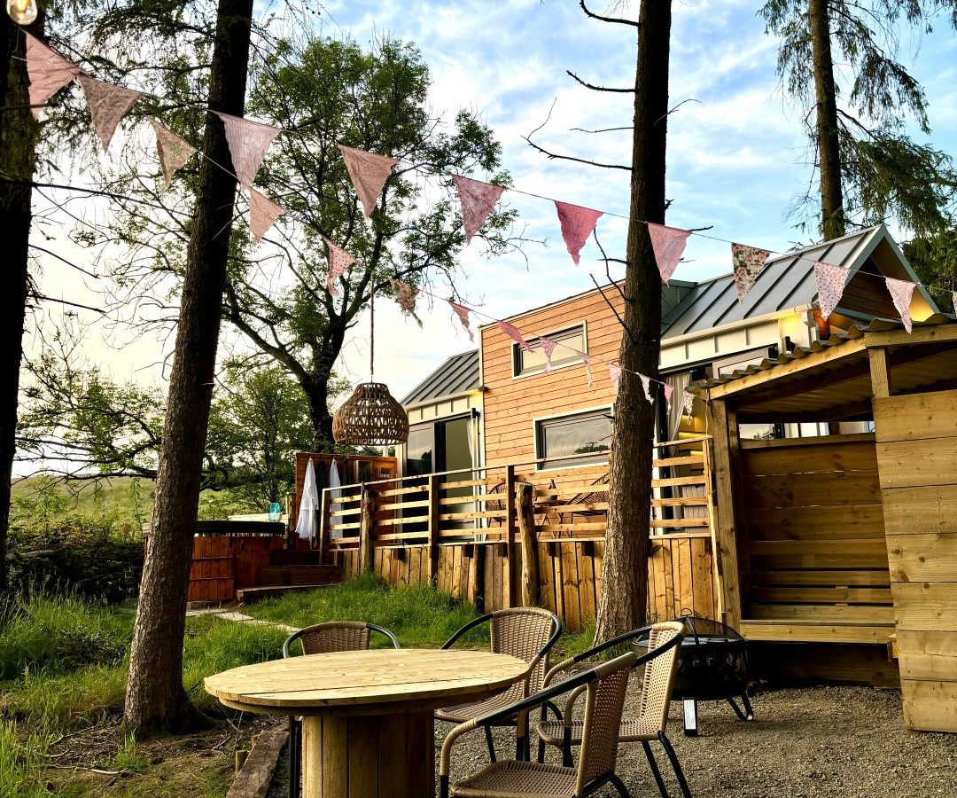 A cozy outdoor seating area with a round wooden table and chairs is nestled in the woods, surrounded by tall trees. A string of colorful bunting is hung above, leading to a modern-design tiny house. The sky is partly cloudy.