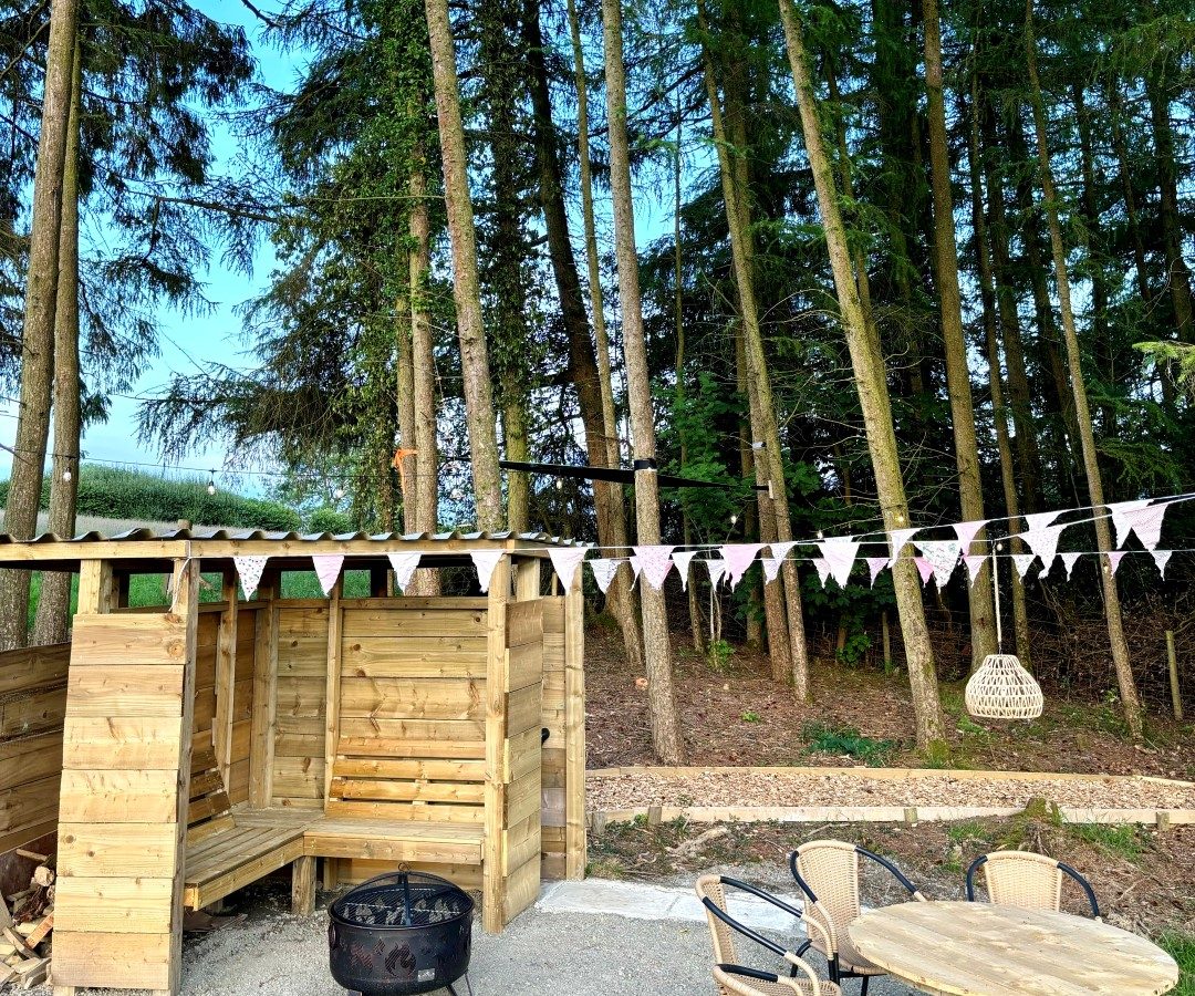 A cozy outdoor seating area beside a tiny house in the woods. The scene features a wooden bench and table set, a fire pit, and triangular string bunting. Tall trees fill the background under a clear blue sky.