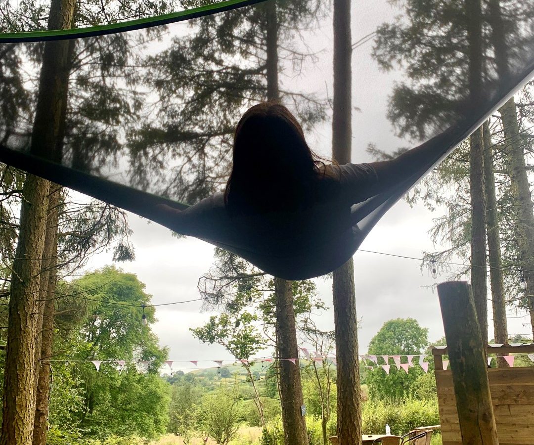 A person relaxes in a black hammock suspended between the trees of the woods. In the background, lush greenery and distant hills unfold beneath a cloudy sky. Nearby, a tiny house peeks through the foliage, while bunting strung between branches adds a festive touch.