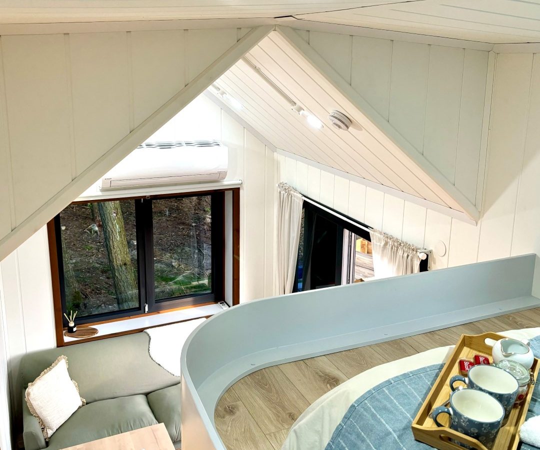 The interior of this tiny house in the woods features a loft bedroom with a neatly made bed and breakfast tray. Below, a cozy living area has a small sofa and dining table by the window. The white walls and ceiling enhance the bright, welcoming space amidst the tranquility of nature.