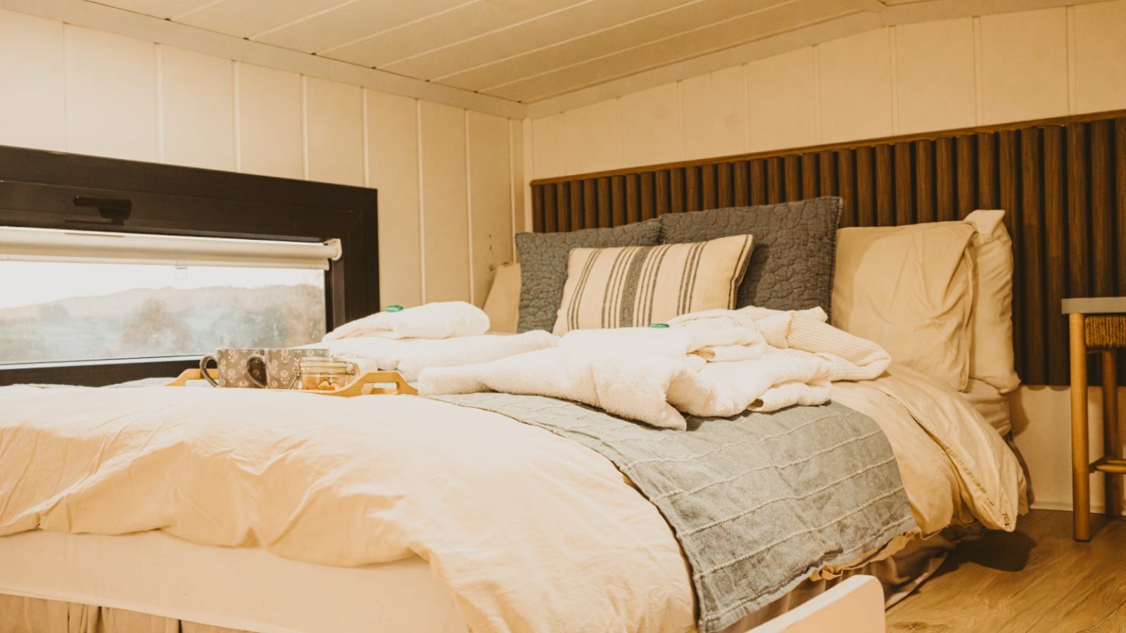 A cozy bed in a well-lit tiny house in the woods with a tray holding cups. The bed is made with pillows and folded blankets, creating a perfect retreat.