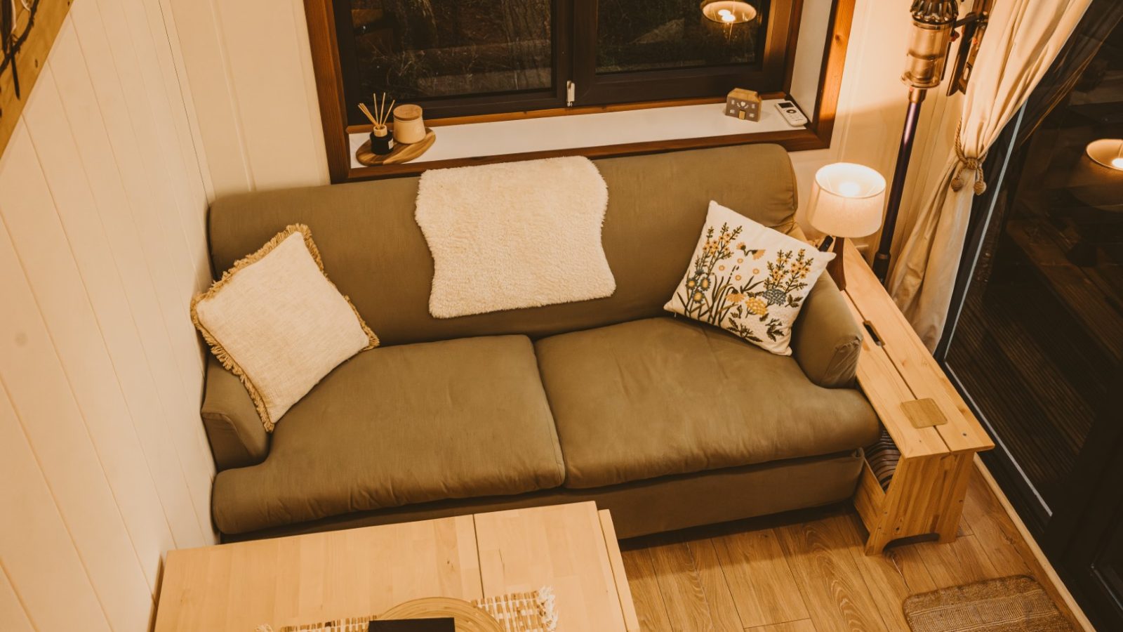 A cozy living room in a tiny house features a beige sofa adorned with decorative pillows, complemented by a wooden table. Warm lighting softly illuminates the space from lamps positioned by the window, creating an inviting retreat amidst the woods.