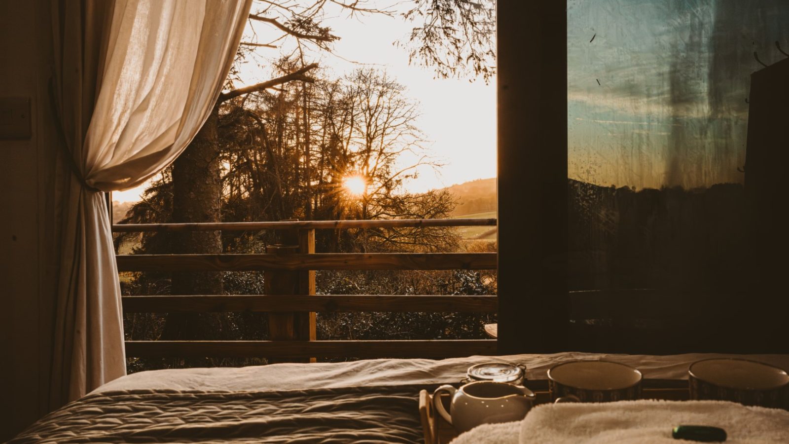 View from a cozy bed in a tiny house, looking out a window at a sunrise over trees and hills, with curtains partially open, nestled in the woods.