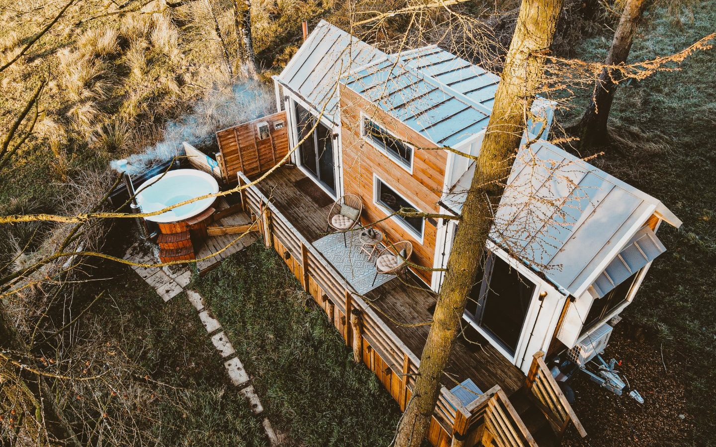 Aerial view of a wooden tiny house nestled in the woods, featuring a deck with a steaming hot tub and two inviting chairs on the porch.