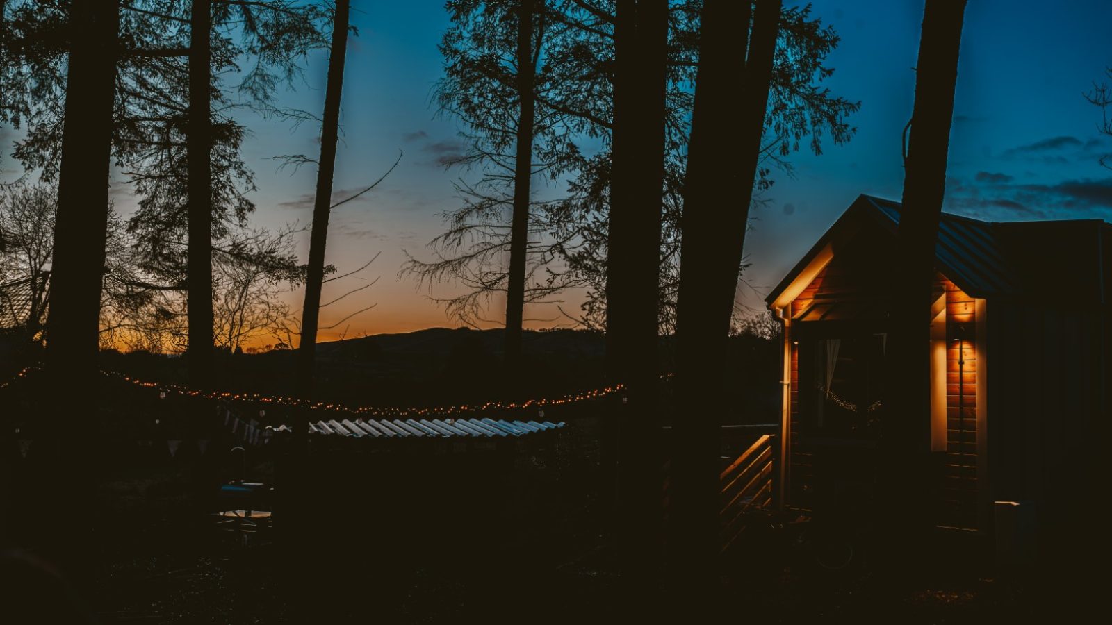 A tiny house with warm lights is nestled among trees at dusk, with a glowing horizon visible beyond.