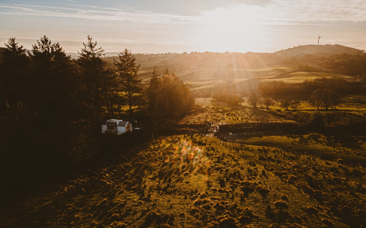 A drone view captures a rural landscape at sunset, featuring rolling hills and a tiny house nestled among the trees beside the woods, with a wind turbine standing tall in the distance.