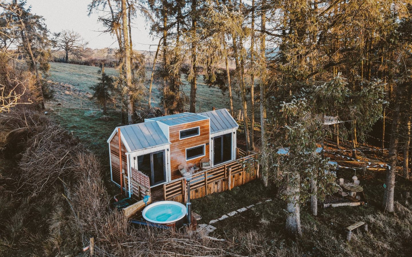         This tiny house in the woods is surrounded by towering trees, with a hot tub outside nestled in the grassy, wooded area.