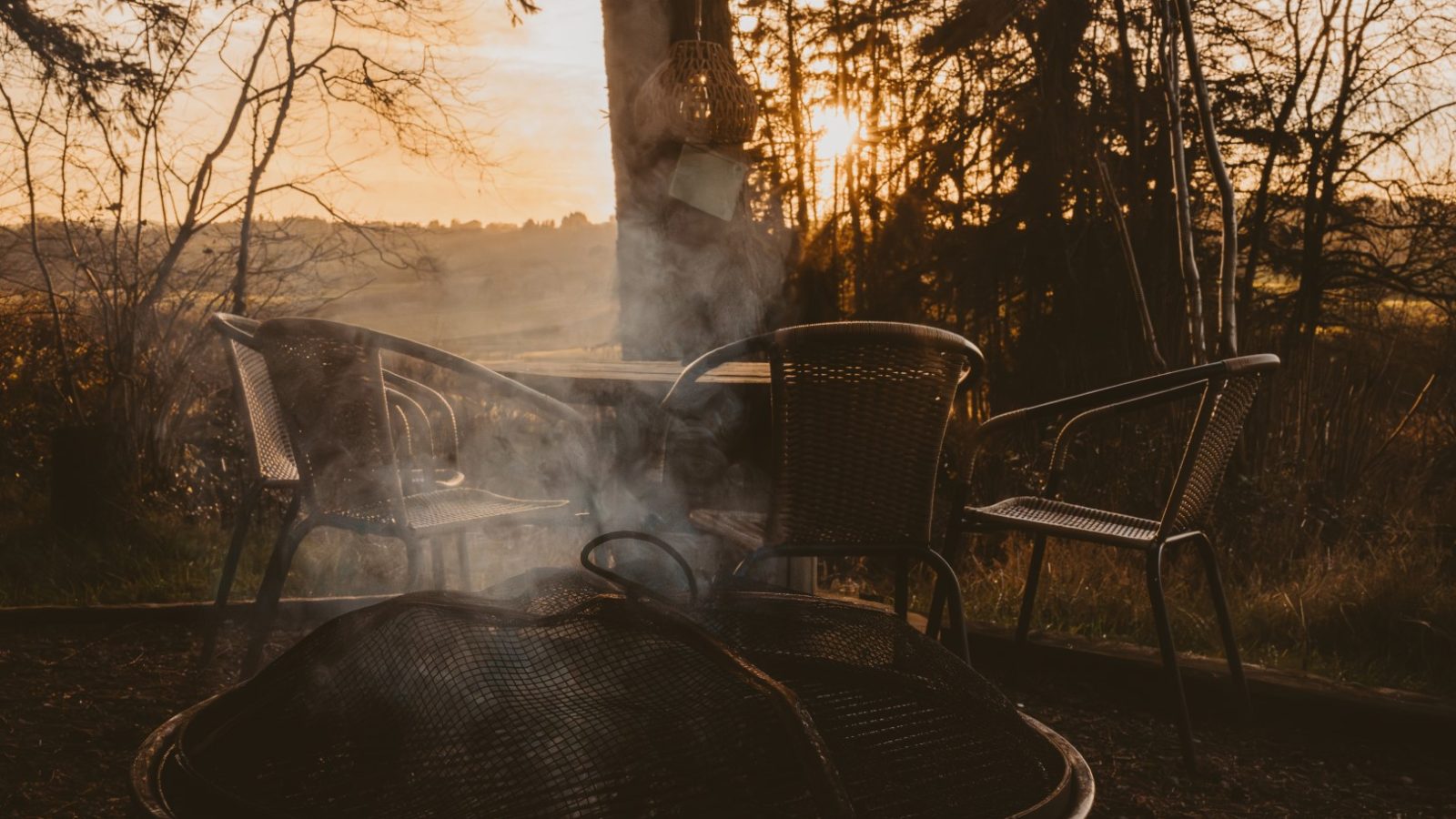 Outdoor seating area with metal chairs and a table near a fire pit, nestled by a tiny house in the woods. Surrounded by trees at sunset, wisps of smoke are visible, creating a cozy ambiance.