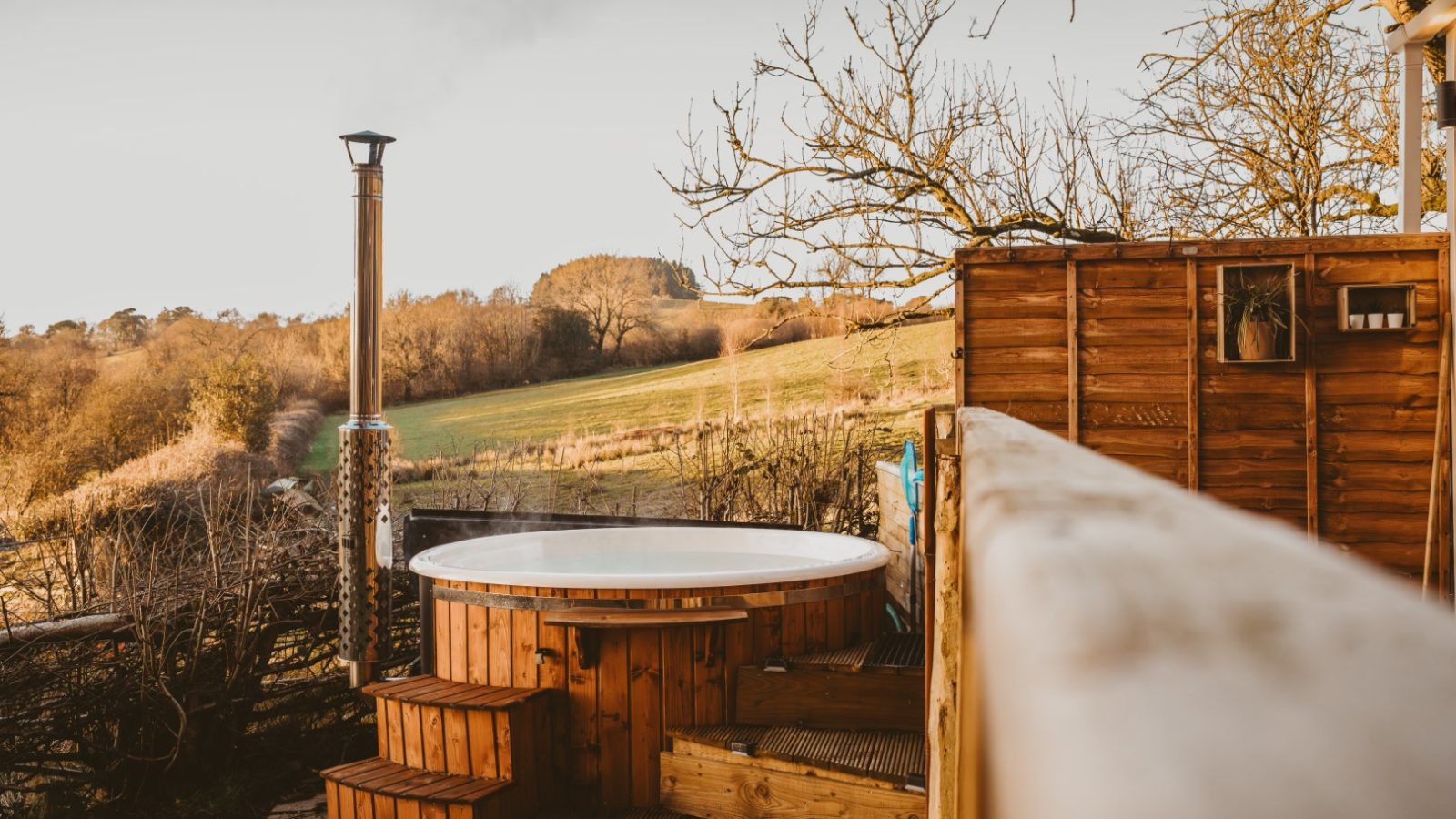 A wooden hot tub on a deck, part of a charming tiny house in the woods, overlooks a scenic rural landscape with trees and fields under a clear sky.