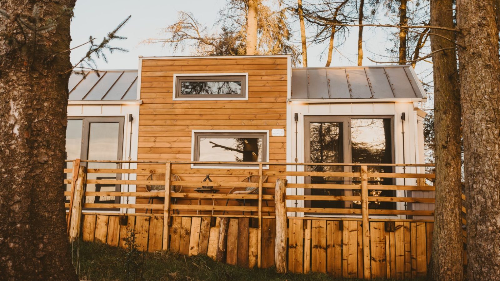 A wooden tiny house with large windows, nestled in the woods among tall trees, and surrounded by a rustic wooden fence.