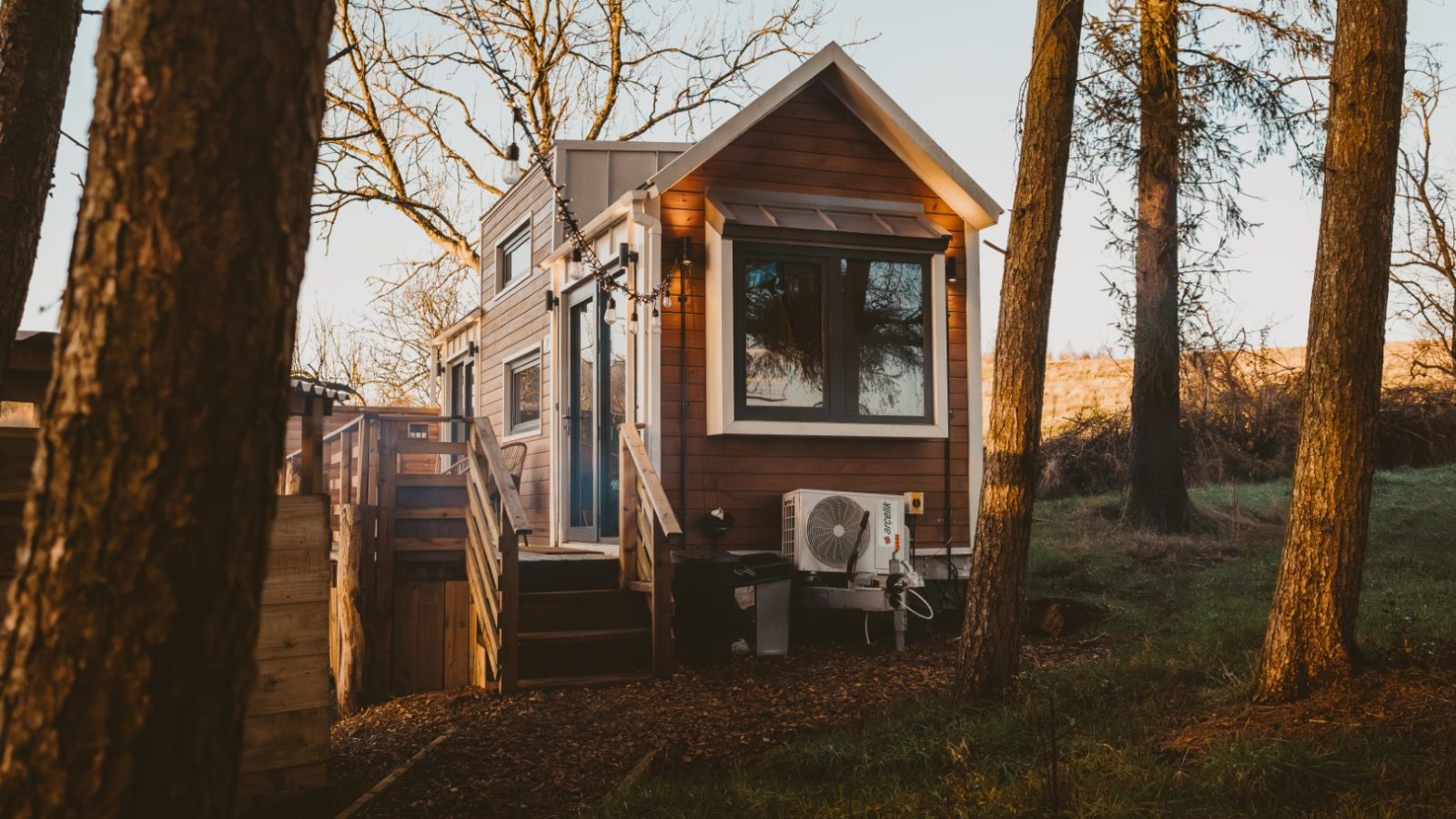 A tiny house nestled in the woods features a cozy porch. Surrounded by tall trees, grass, and fallen leaves, it basks under a clear sky.