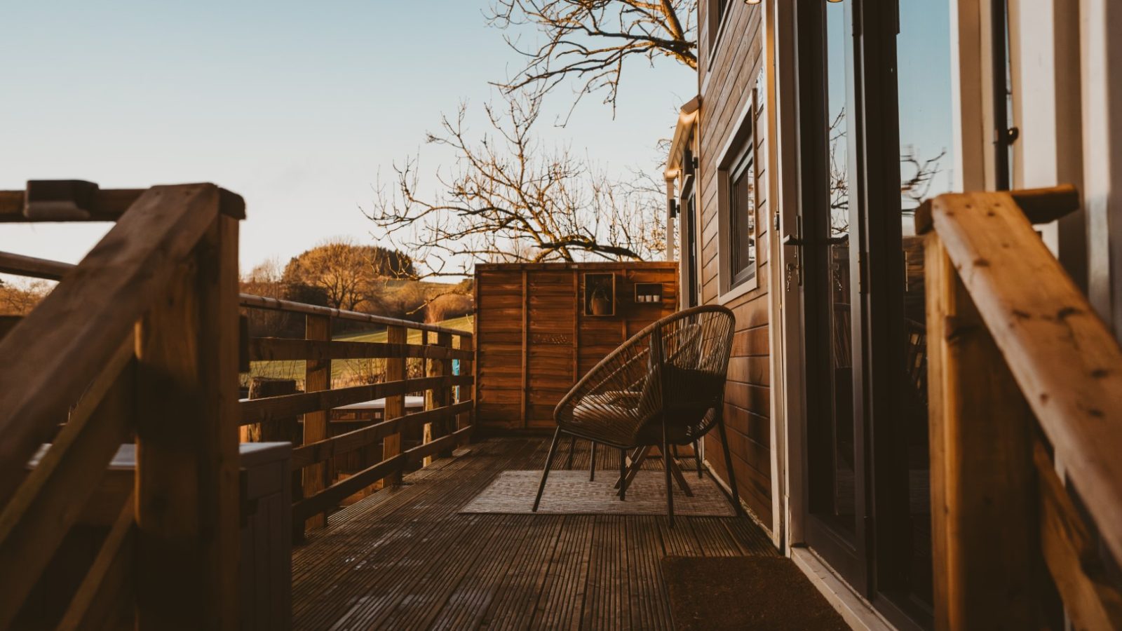 A charming wooden deck with railing and wicker chair offers a serene escape, overlooking sprawling fields and dense woods under a clear sky, making it the perfect retreat for your tiny house adventure.