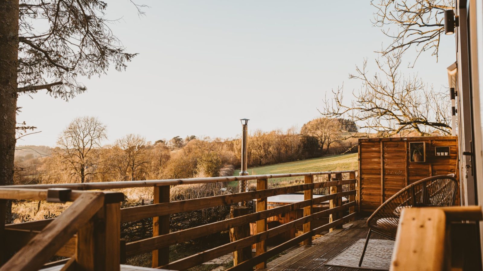 A wooden deck attached to a cozy tiny house in the woods overlooks a grassy field and trees, all under a clear sky and bathed in warm lighting.