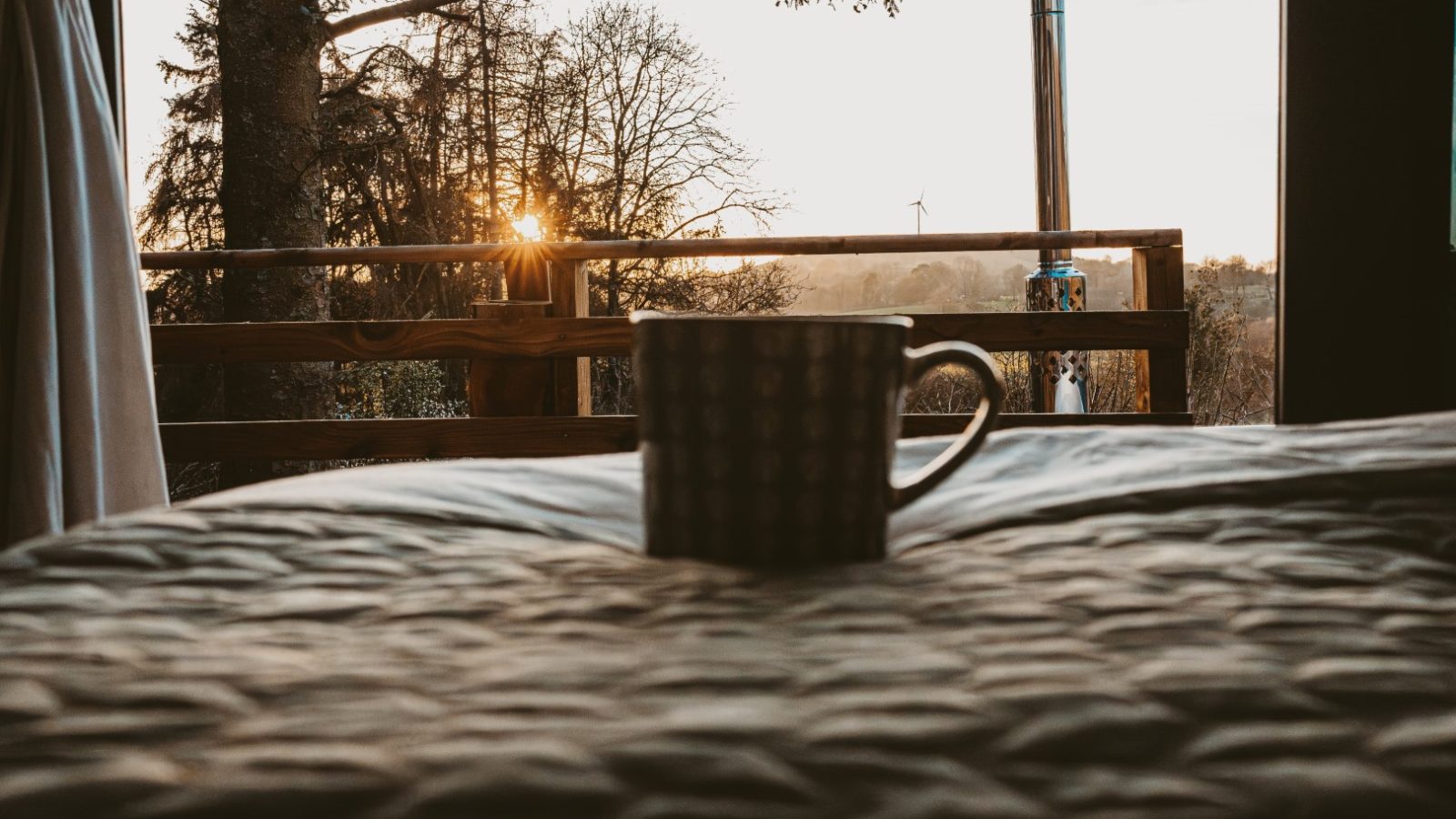 A mug rests on a bed in a cozy tiny house, offering a sunlit view of the balcony and towering woods just beyond the window.