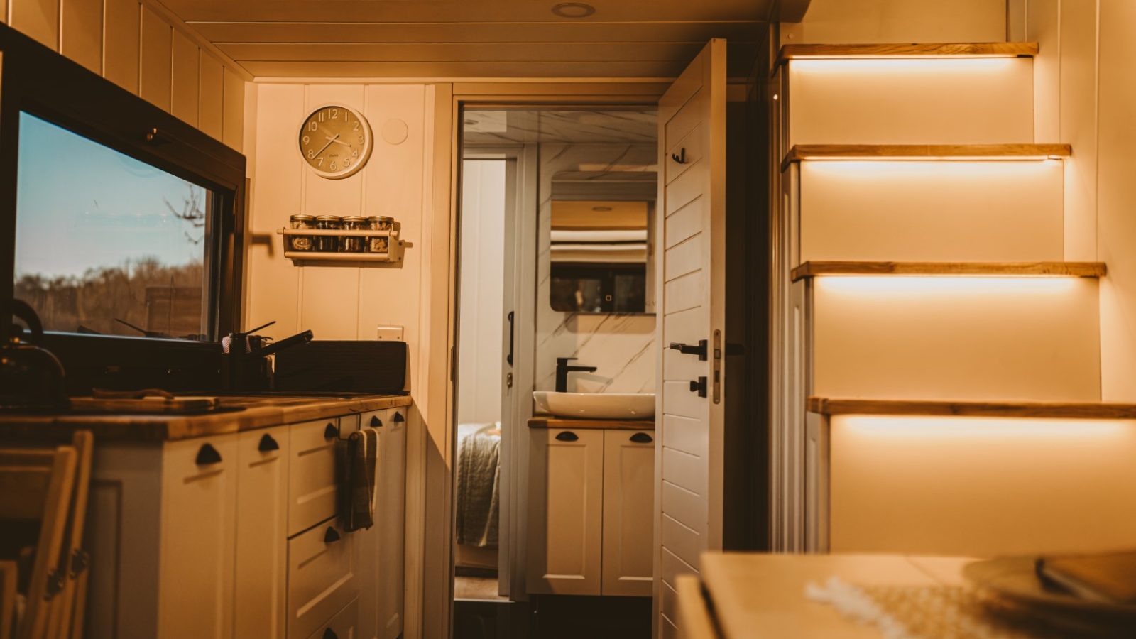 The interior of this cozy, well-lit tiny house camper features a kitchen, staircase, and a view into the bedroom. Warm woods and white accents create a charming and inviting atmosphere throughout.