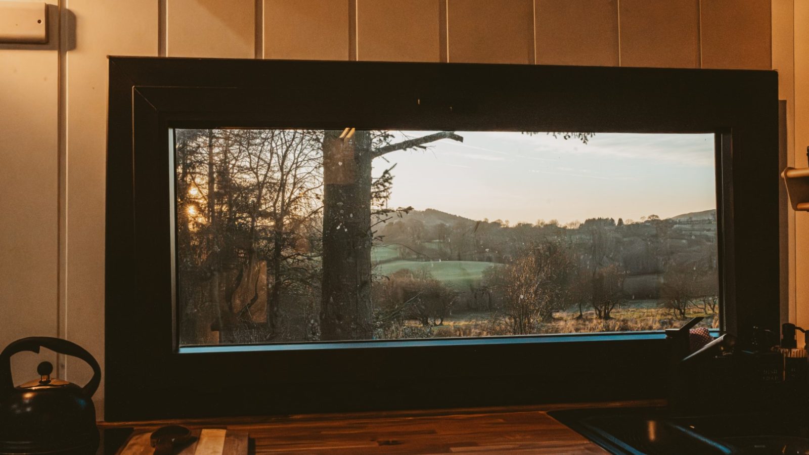 View through a large kitchen window of a landscape at sunset, with trees and distant hills embracing the tiny house. The kitchen counter is visible in the foreground, offering a cozy scene.