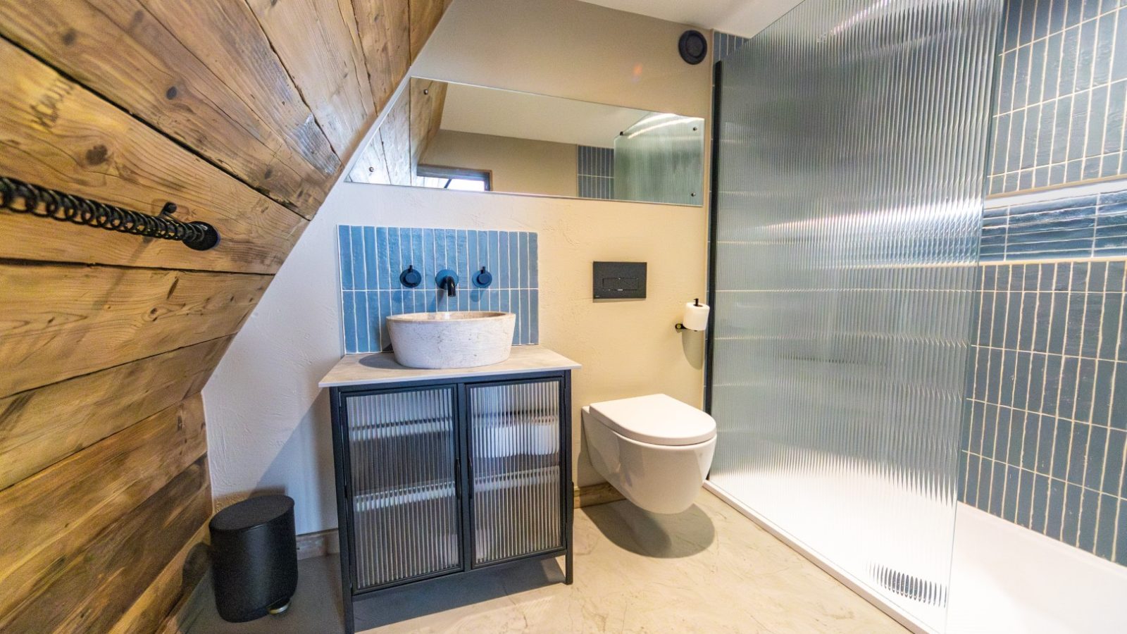 A modern bathroom with a sloped wooden ceiling. The space features a floating toilet, a vanity with a round stone sink, and a large mirror above it. The glass-enclosed shower boasts blue tile accents reminiscent of Tri-Pod designs. The floor is tiled in a light color.