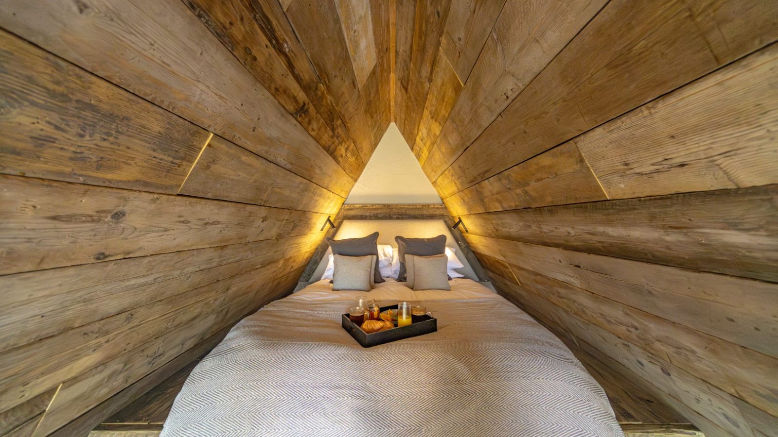 A cozy bedroom with wooden plank walls forming a triangular shape at the head of the bed. The neatly made bed with grey bedding and two pillows features a black tray with a breakfast arrangement, including a croissant and drinks, resembling the balanced setup of a tri-pod.