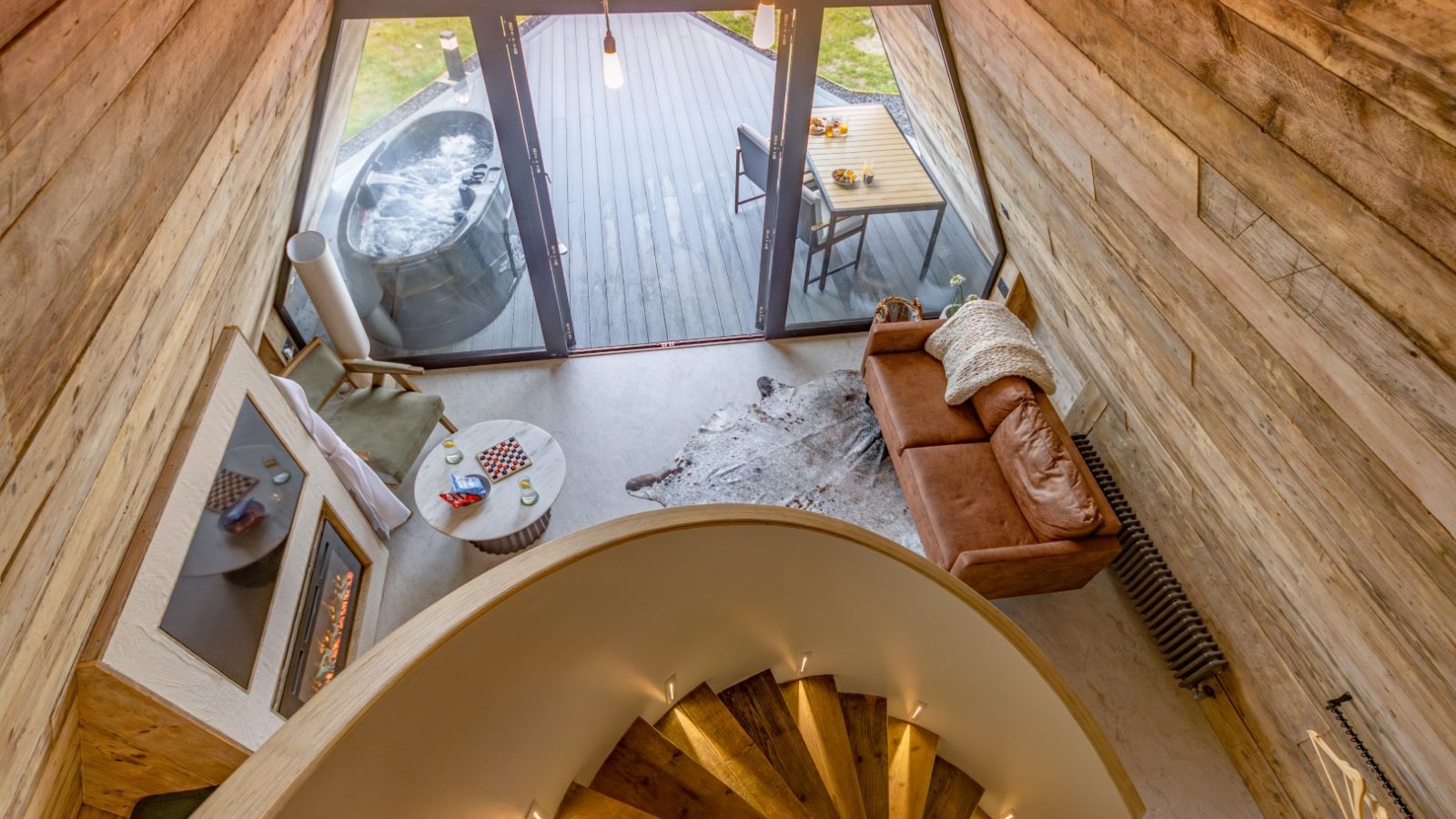 This image captures a cozy, modern living space with wooden walls and a spiral staircase. A brown leather sofa faces a fireplace, and a large window reveals a wooden deck with a hot tub beside some photography equipment, including a tri-pod. A small table with two chairs is set near the hot tub.