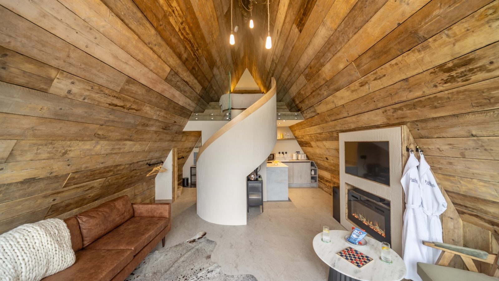 A cozy, modern loft with an A-frame wooden interior. It features a brown leather sofa, a spiral staircase, a fireplace, a small round table with checkers, and a tripod for photography enthusiasts. The kitchen is visible in the background, and robes hang by the fireplace.