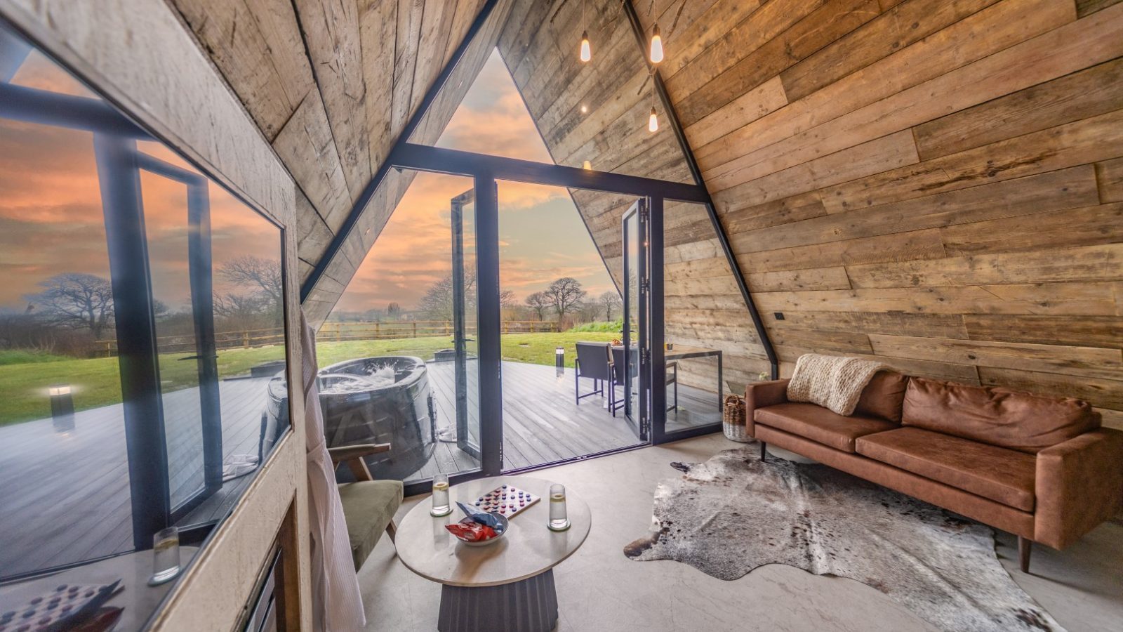 A cozy A-frame cabin interior features wooden walls, a brown sofa, and a modern coffee table with a chessboard. Large glass doors open to a deck with a hot tub, tripod set up for photography, and outdoor dining area, overlooking a scenic sunset with trees and a grassy landscape in the background.