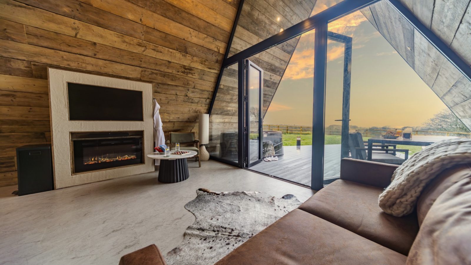 A cozy, modern cabin interior with wooden walls and large floor-to-ceiling windows providing a scenic view of the outdoors. The room features a fireplace, a small coffee table, a cowhide rug, and a brown leather sofa with a blanket. A tripod stands ready on the outside deck visible through the sliding doors.