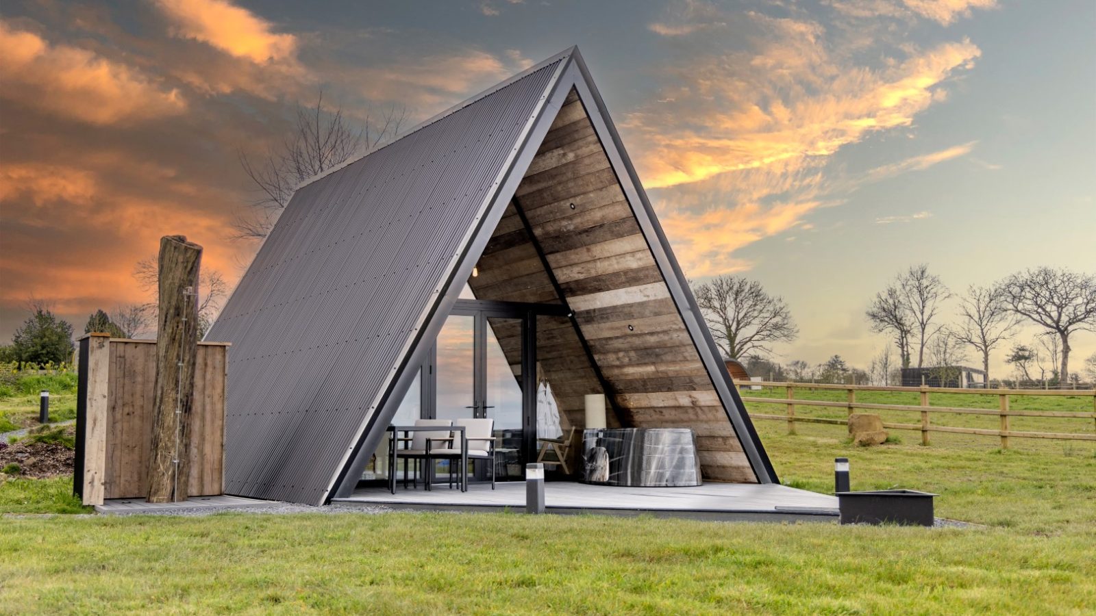 A modern A-frame cabin with large glass windows is captured beautifully under a dramatic, colorful sunset sky. The cabin features a small patio area with outdoor seating and sits in a grassy, fenced field with a few bare trees—a picture-perfect spot where your tripod stand can capture stunning views.