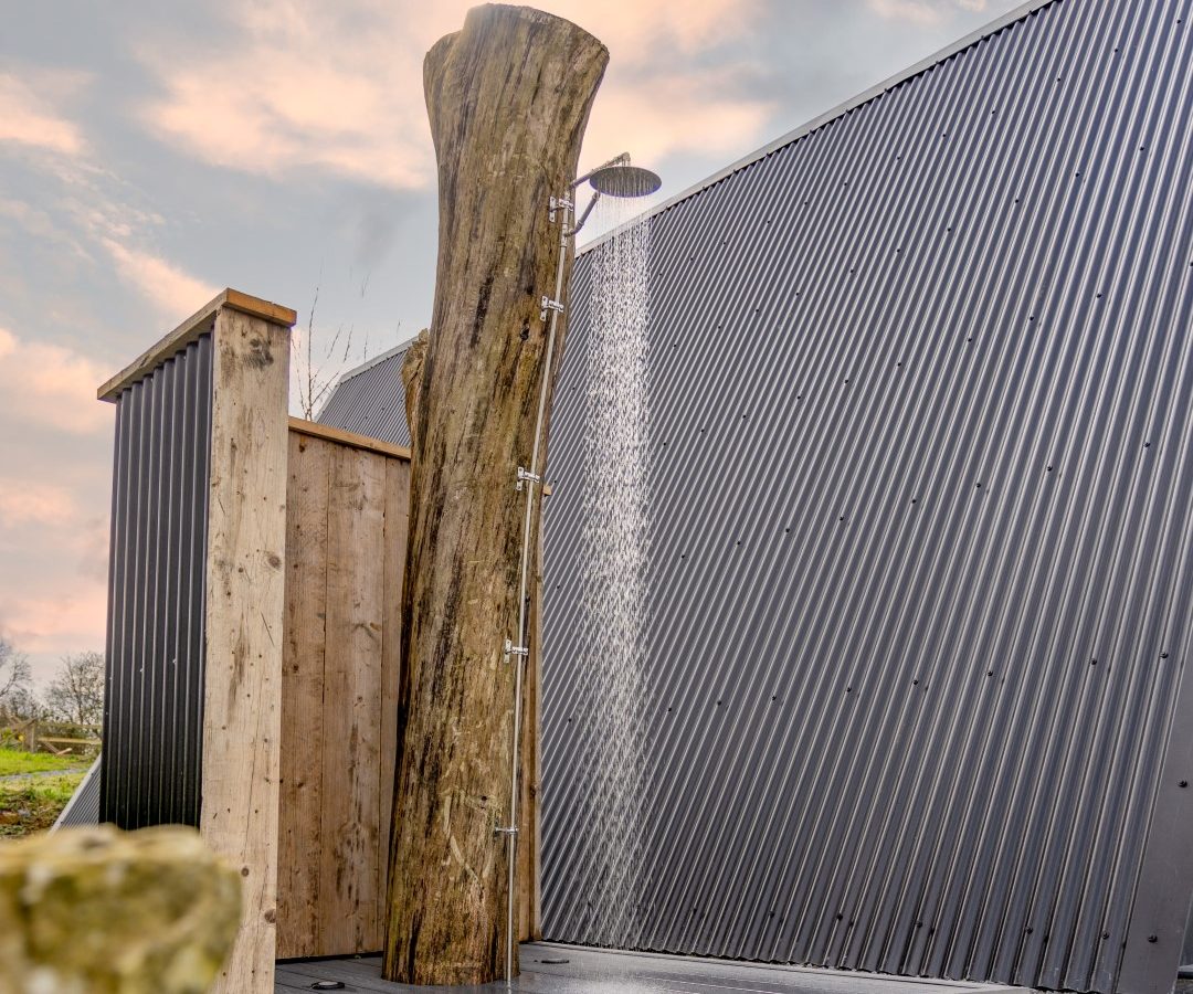 An outdoor shower features a large, vertical log as the shower column against a corrugated metal wall. With Tri-Pod in hand, capture water flowing from the shower head, the gravel pathway, and grass in the foreground—all set under a pink evening sky.