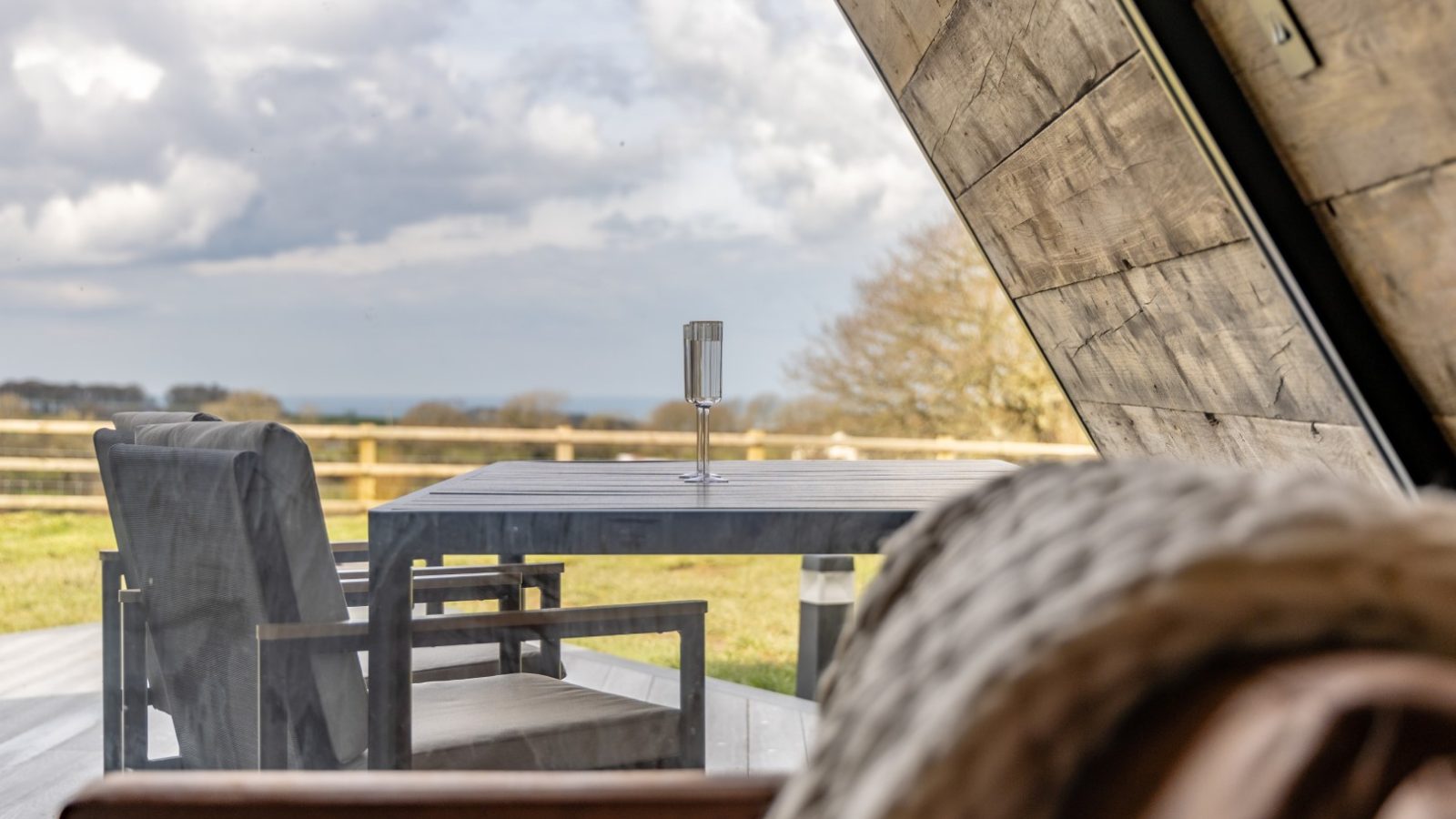 A cozy, modern cabin interior with a view through a large window of an outdoor patio featuring a dining table and chairs. The patio overlooks an expansive, grassy landscape under a partly cloudy sky. Inside the cabin, near the textured blanket draped over a surface, stands a sleek tri-pod lamp.