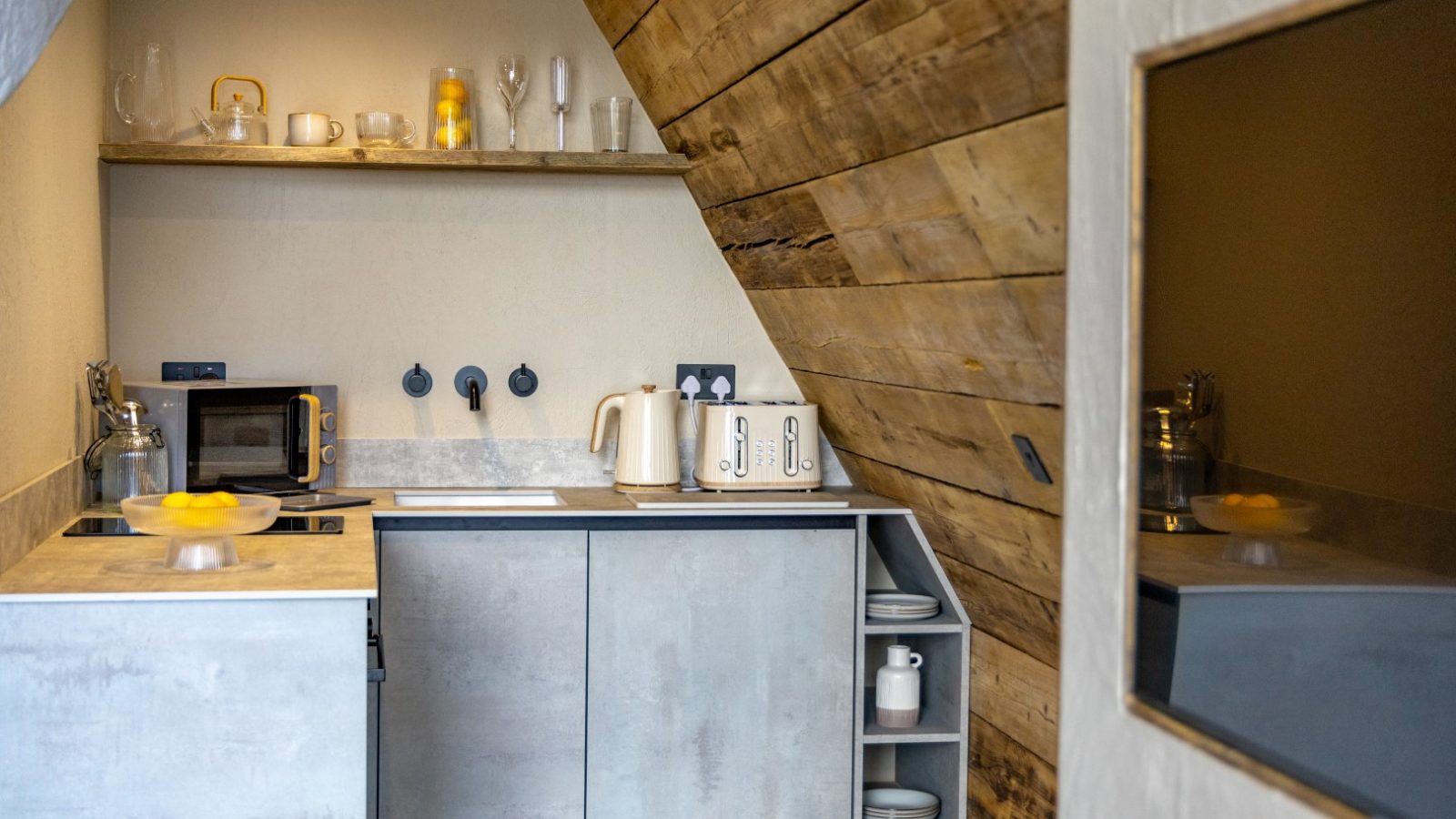 A small, modern kitchen with rustic wooden walls showcases grey cabinets and a sleek electric stovetop akin to a precision instrument. Tri-Pod inspired shelving holds glassware and a bowl of lemons. A kettle graces the counter, while the window reflects the thoughtfully curated decor.