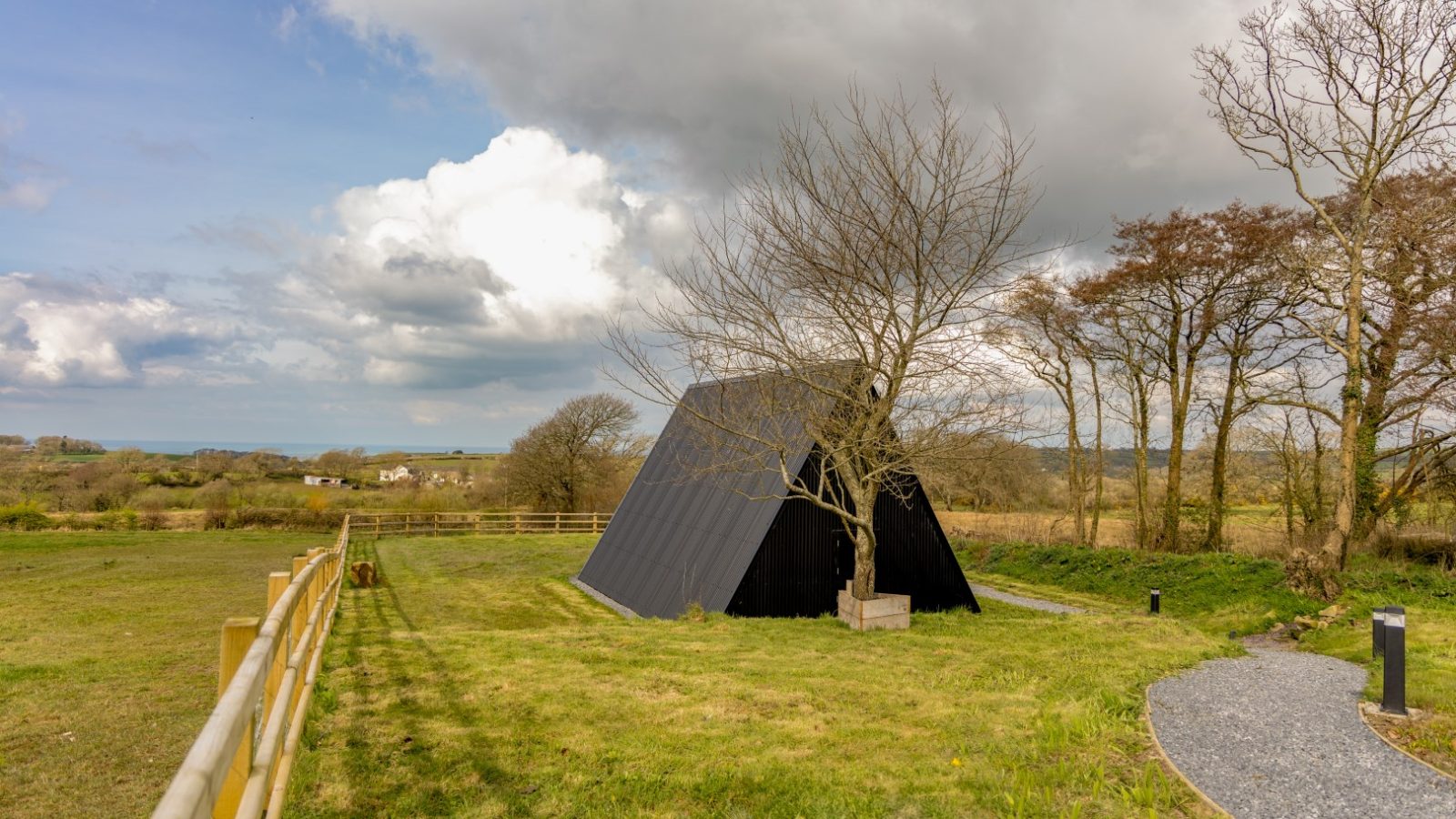 A modern, black A-frame cabin stands in an open field under a cloudy sky. Nearby, a leafless tree complements the scene. The path leading to the cabin is perfect for capturing with photography equipment like a sturdy tripod. Sparse trees and distant houses complete this picturesque setting.