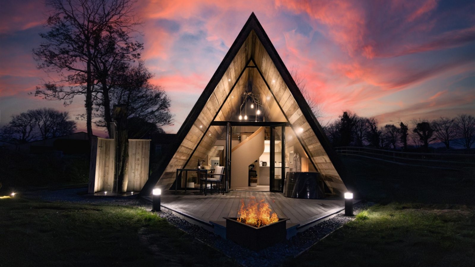 A-frame Tri-Pod cabin with an illuminated interior, set against a vibrant sunset sky. The triangular structure features large glass doors, warm lights, and a firepit on a wooden deck. Silhouetted trees and a grassy landscape surround the cabin.