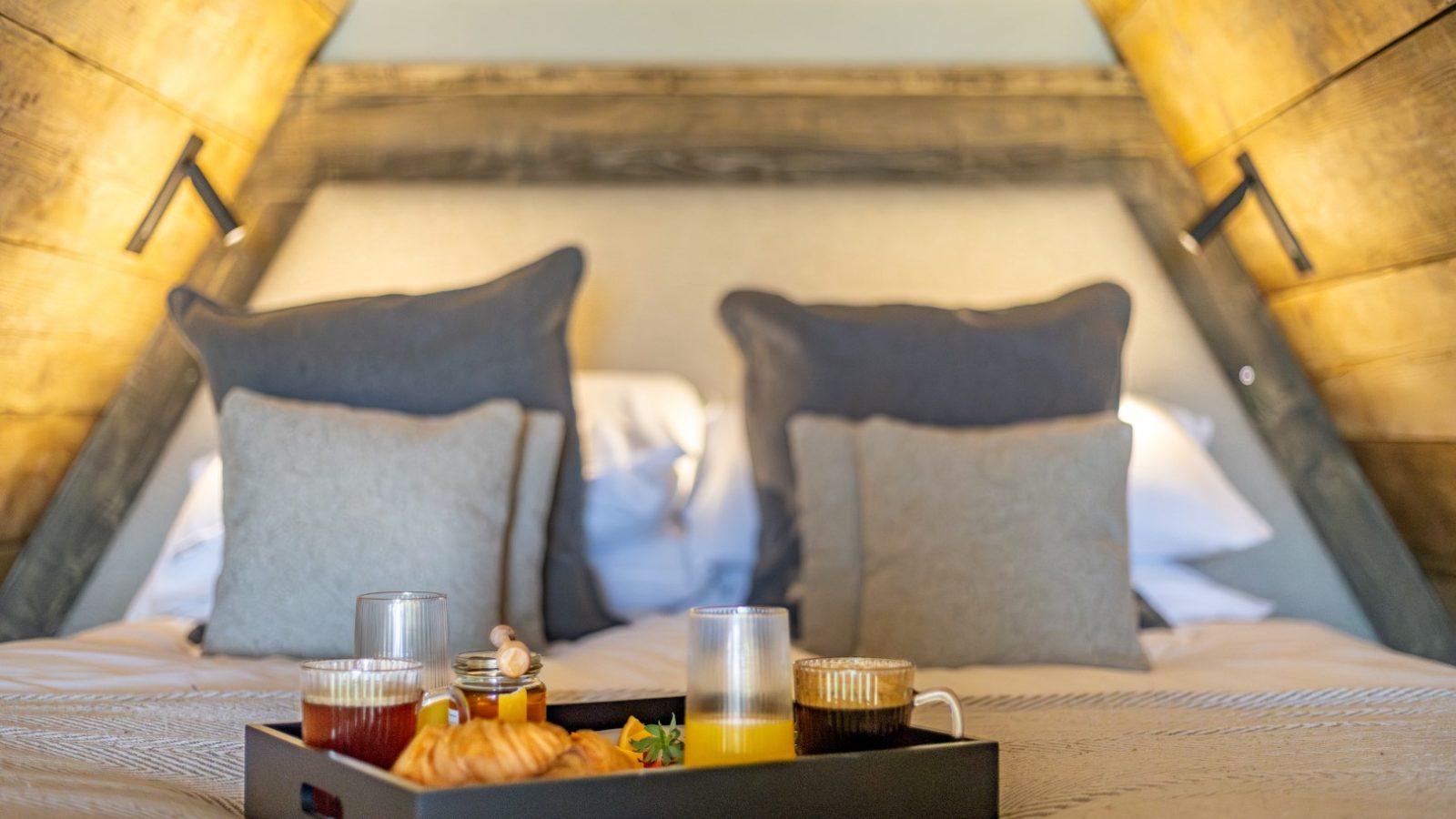 A cozy, wooden-accented bedroom features a neatly made bed with two large pillows and two smaller ones propped against the headboard. A tray with croissants, drinks, and breakfast items sits atop the bed, bathed in soft, warm light from a cleverly positioned Tri-Pod lamp.