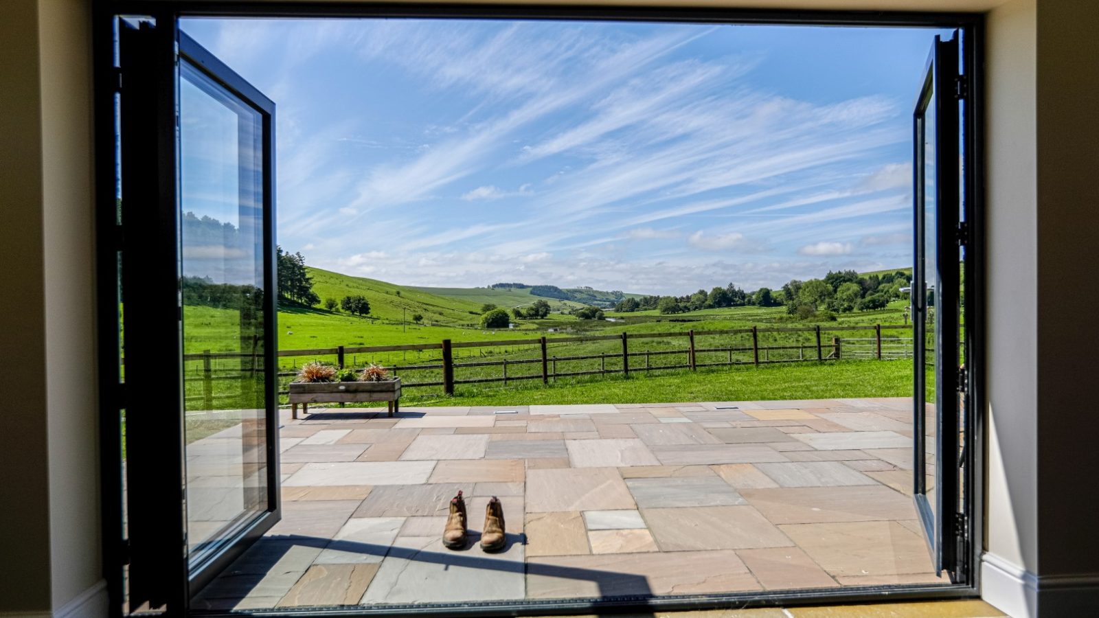 A pair of brown boots sits on a stone-tiled patio in front of open glass doors with black frames at Waen Farm House. The doors lead out to a picturesque green countryside under a blue sky with wispy clouds. A wooden bench with flower pots is visible to the left.