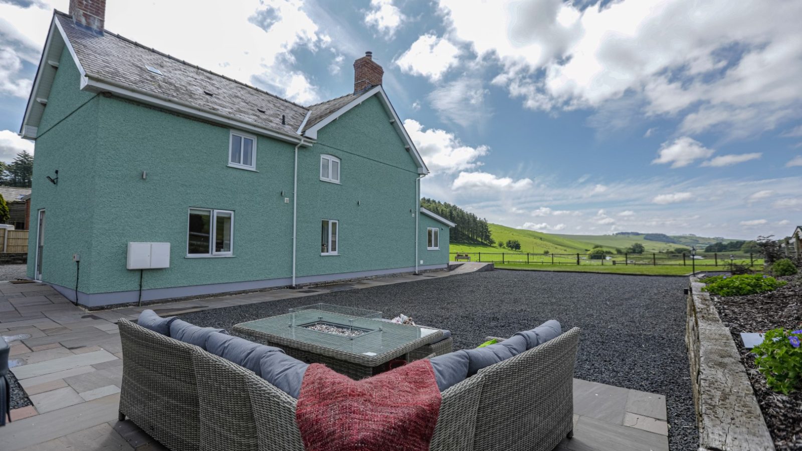 A two-story green house, reminiscent of a charming Waen Farm House, boasts a grey roof and is surrounded by a spacious patio area. The patio features a grey outdoor sectional sofa with a red blanket, set around a fire pit. In the background, rolling green hills stretch under a partly cloudy sky.