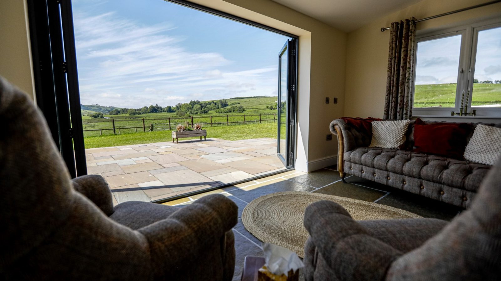 A cozy farmhouse living room with two gray armchairs faces large, open folding glass doors that lead to a sunny patio. The patio overlooks a serene countryside with green fields and hills under a blue, lightly clouded sky. A round rug lies between the chairs, adding a touch of warmth.